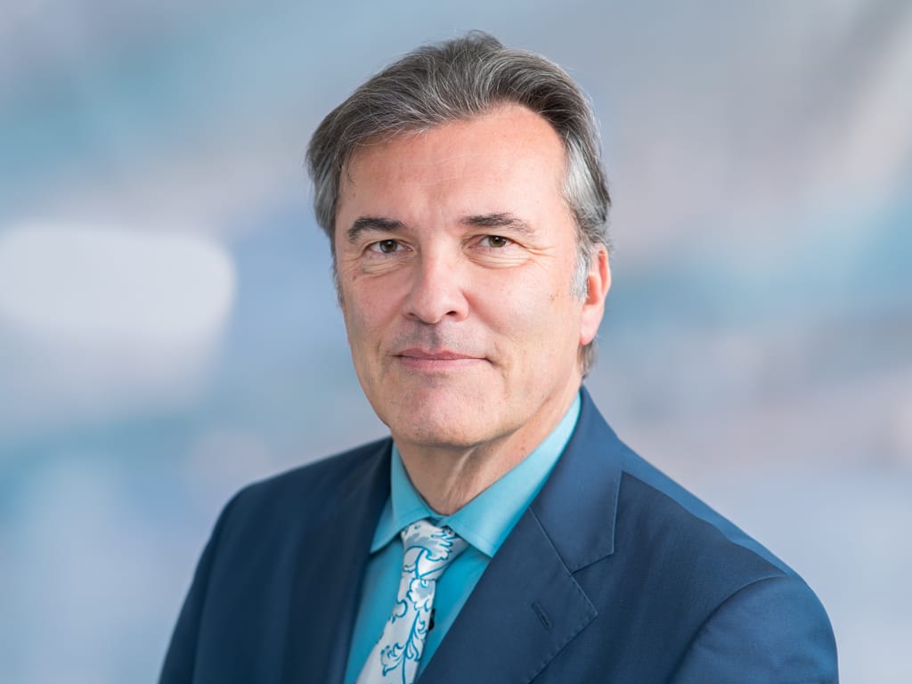 Man wearing a blue suit jacket, turquoise shirt, and patterned tie against a blurred background