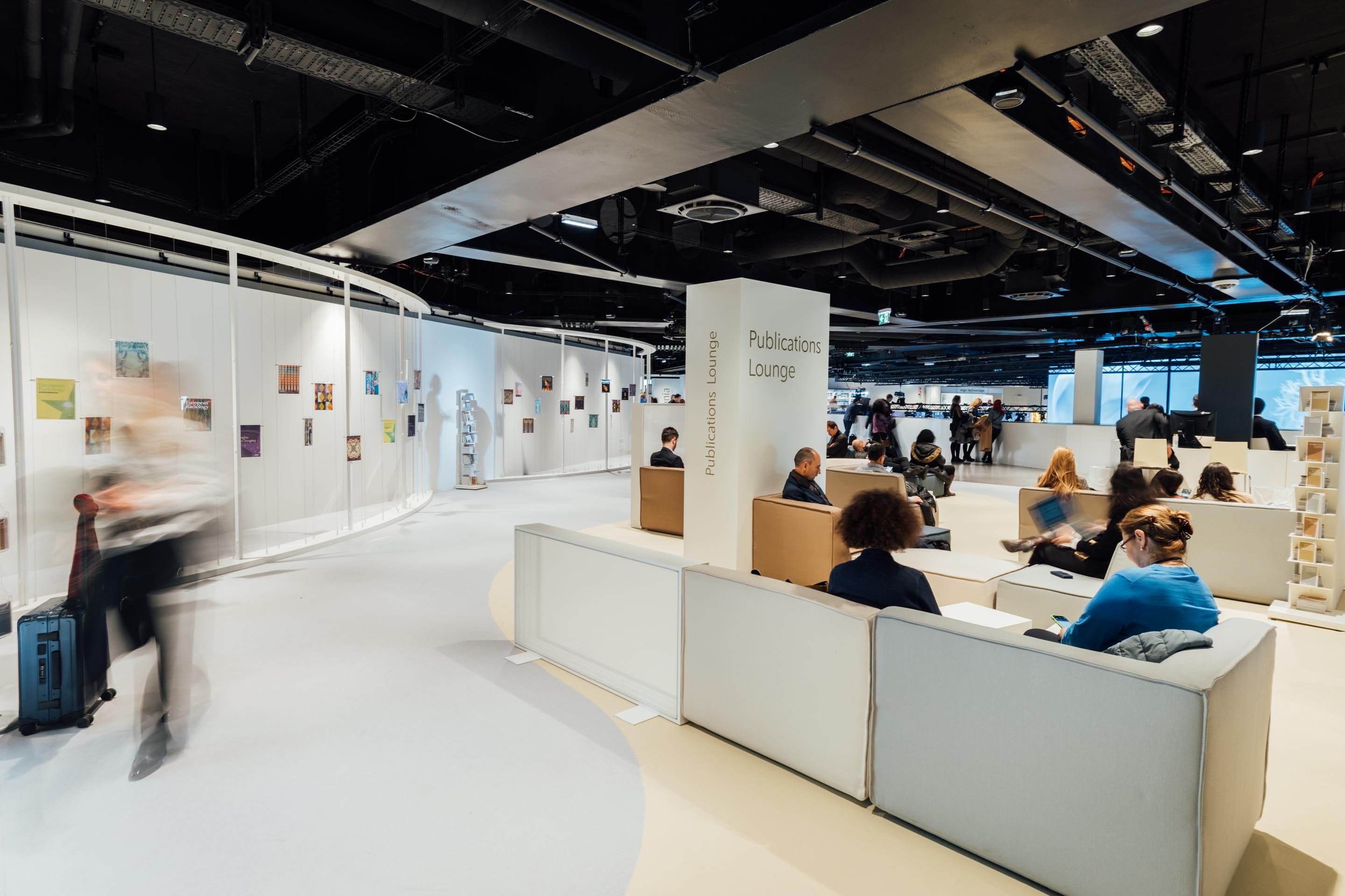 Innenraum mit Sitzbereich und Säule mit Schrift „Publications Lounge“, Personen sitzen und gehen vorbei