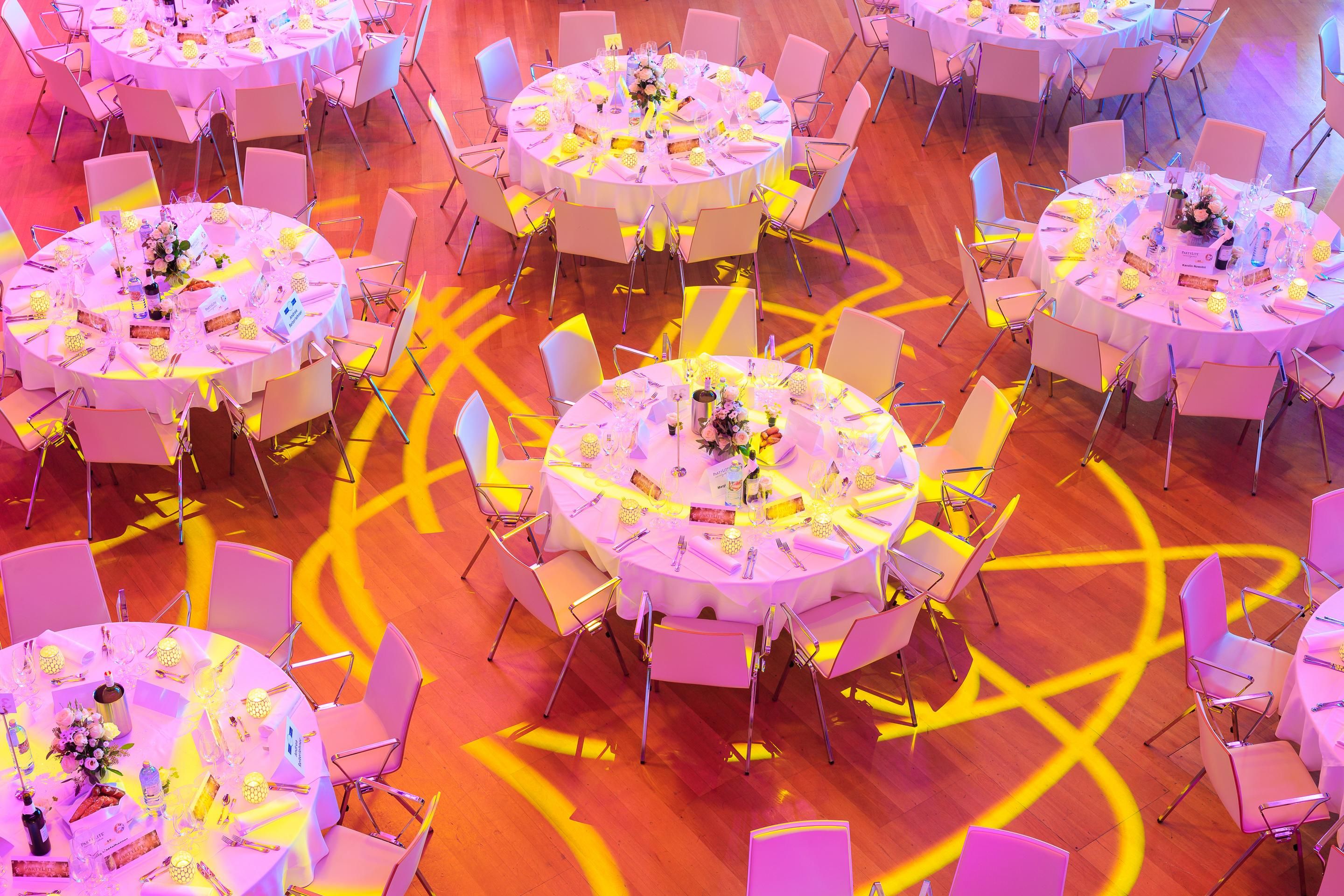 Round banquet tables with white tablecloths and chairs under yellow and pink lighting patterns on wooden floor