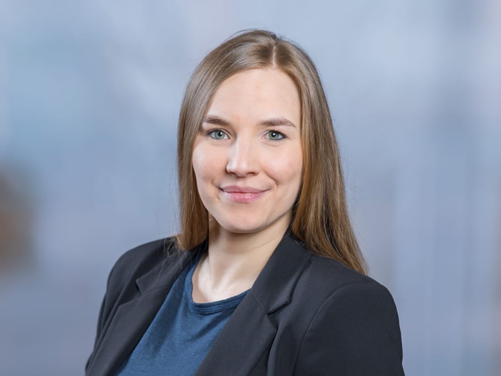 Person with long light brown hair wearing a dark blazer and blue shirt against a blurred background