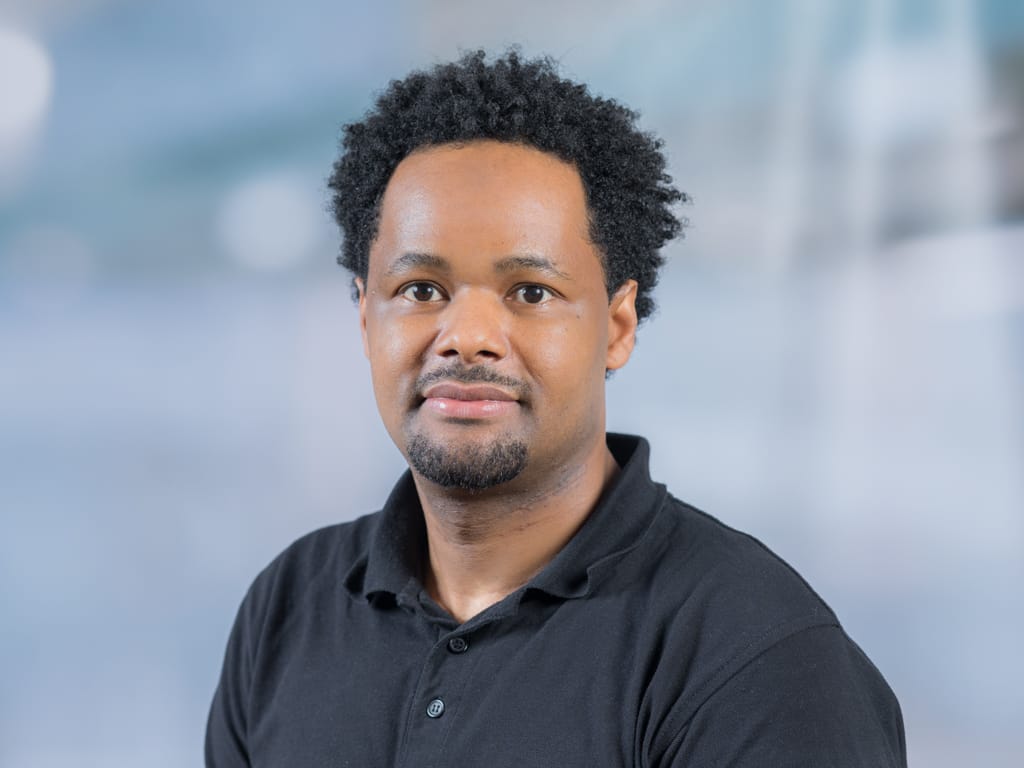 Person with short curly hair wearing a black collared shirt against a blurred background