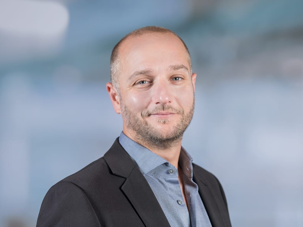 Man wearing a black blazer and blue patterned shirt against a blurred background