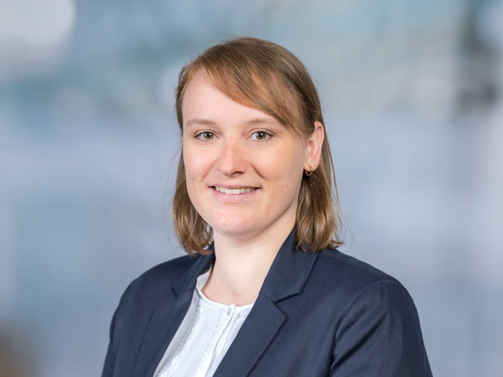 Person mit schulterlangen braunen Haaren, weißer Bluse und dunkelblauem Blazer vor unscharfem Hintergrund