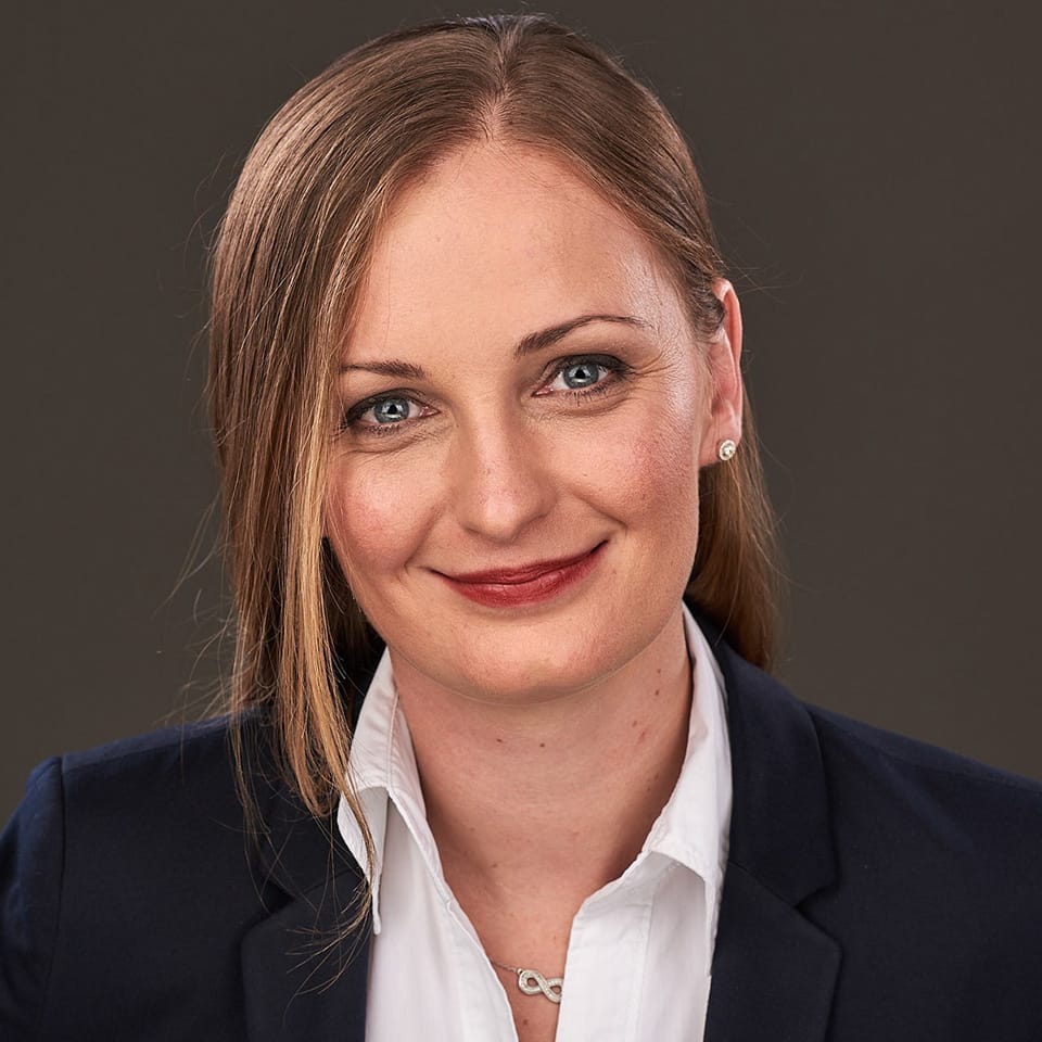 Person mit langem Haar, weißem Hemd, dunklem Blazer und Schmuck am Hals und Ohr.