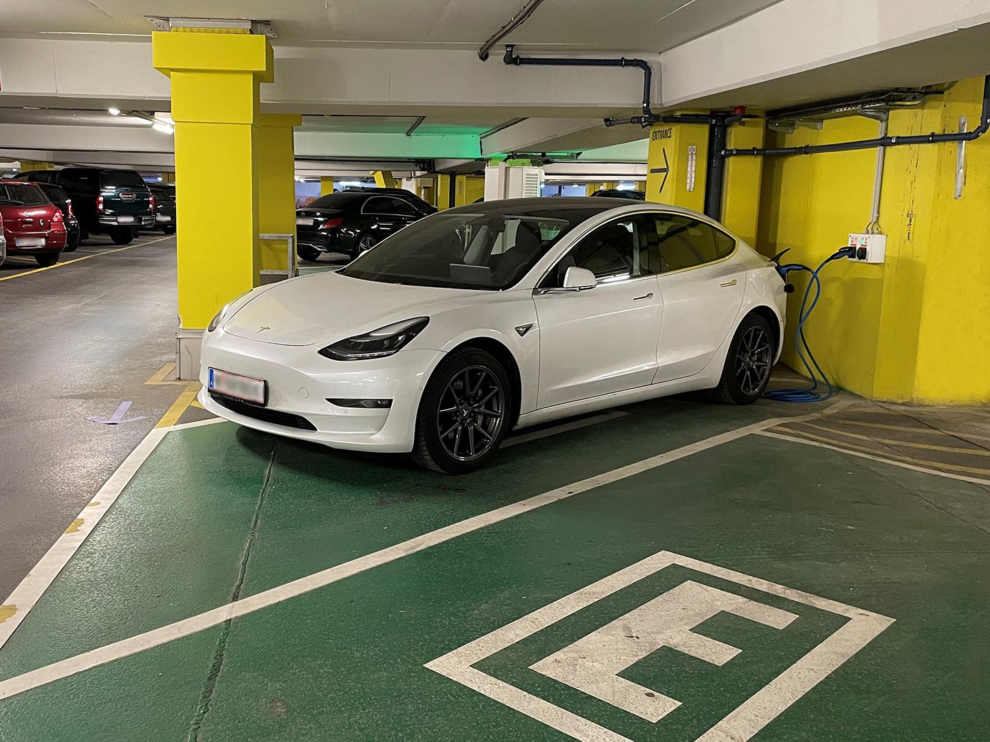 Weißes Elektroauto lädt an Ladestation in Tiefgarage mit gelben Säulen und grünem Boden.