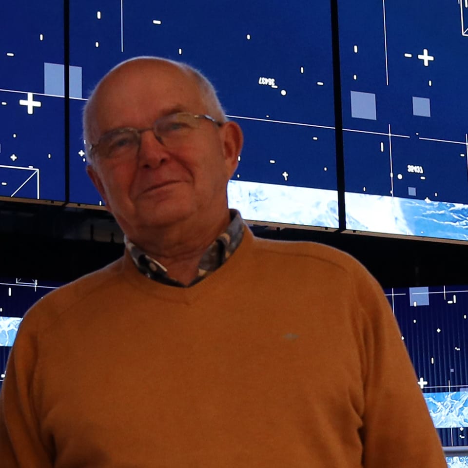 Mann mit orangefarbenem Pullover vor blauem Bildschirm mit weißen Symbolen und Zahlen