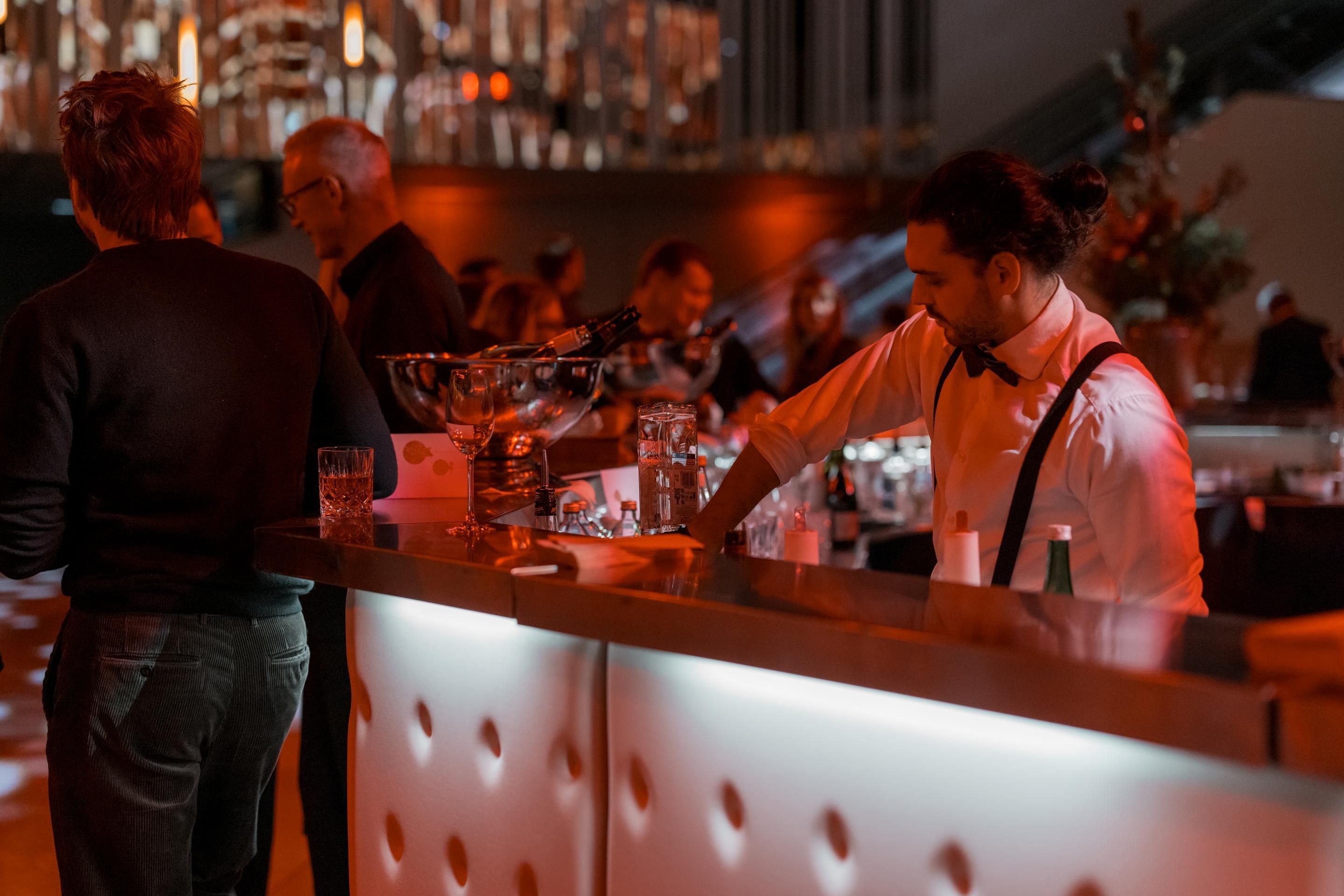 Bar mit beleuchtetem Tresen, Barkeeper in weißem Hemd und schwarzer Fliege, Gäste im Hintergrund