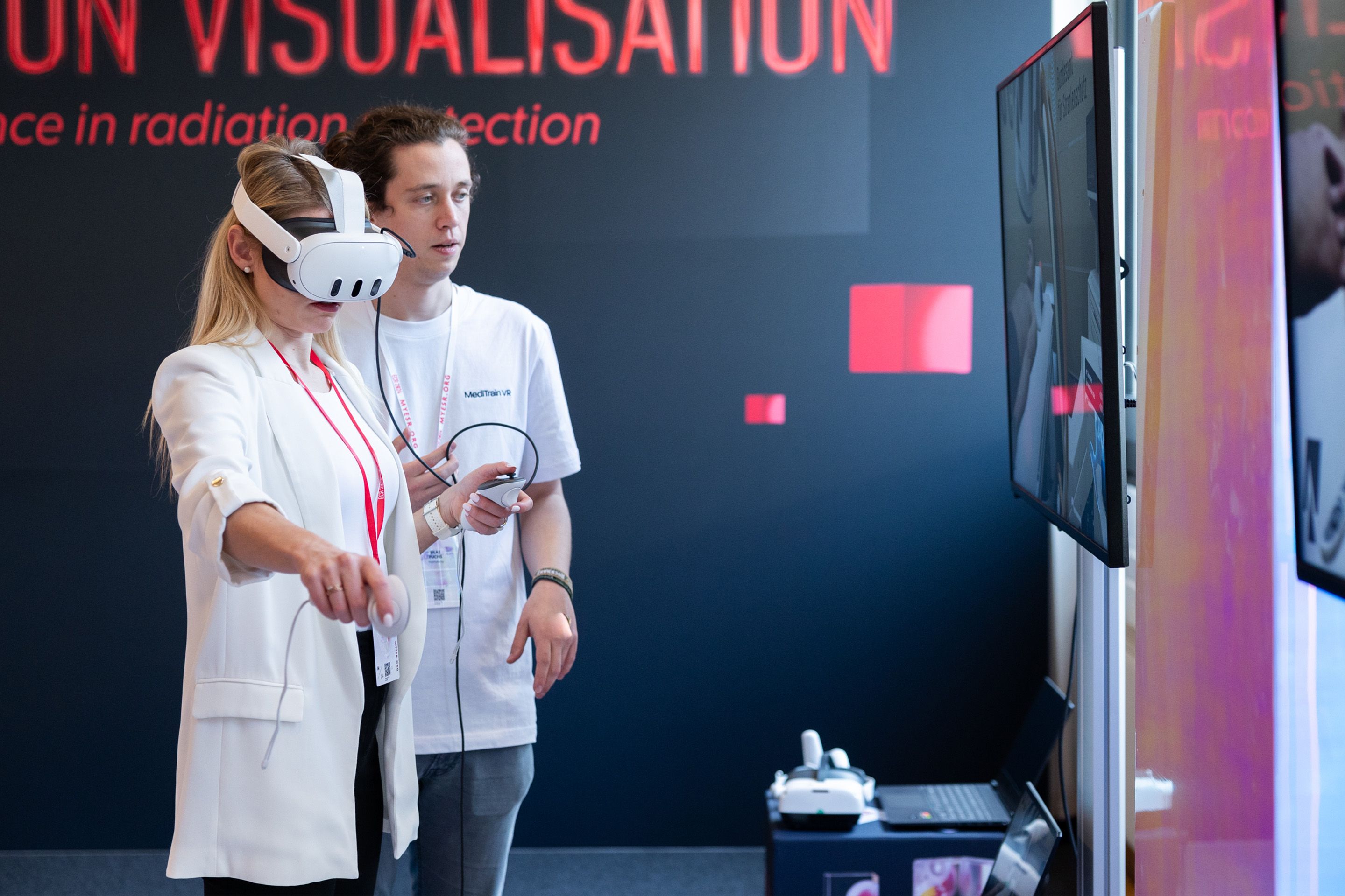 Woman wearing virtual reality headset and holding controllers, man standing behind her.