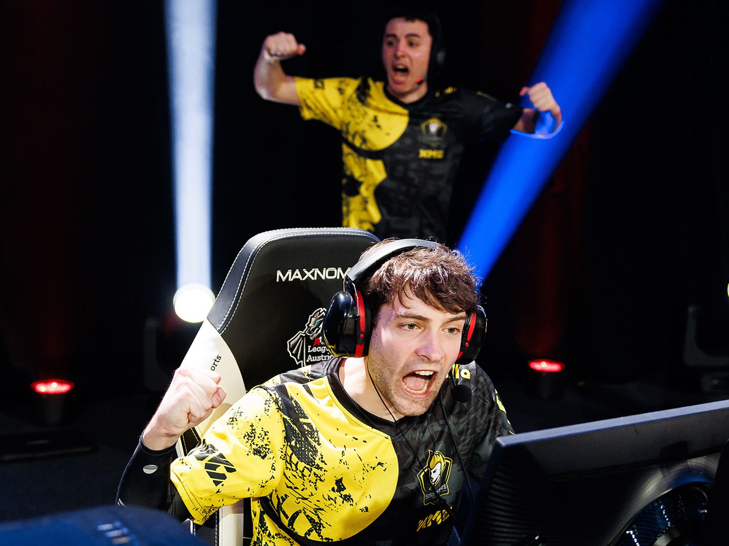 Two people in yellow and black esports jerseys celebrating in front of a gaming setup with headset and monitor.