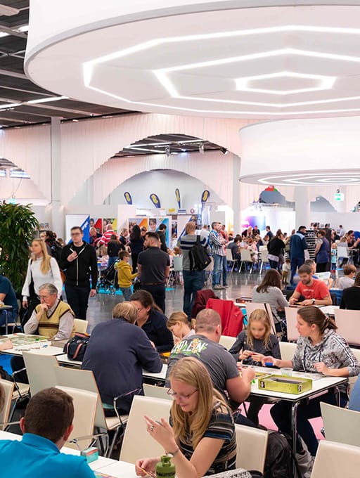Viele Menschen sitzen an Tischen und spielen Gesellschaftsspiele in einer großen Halle.