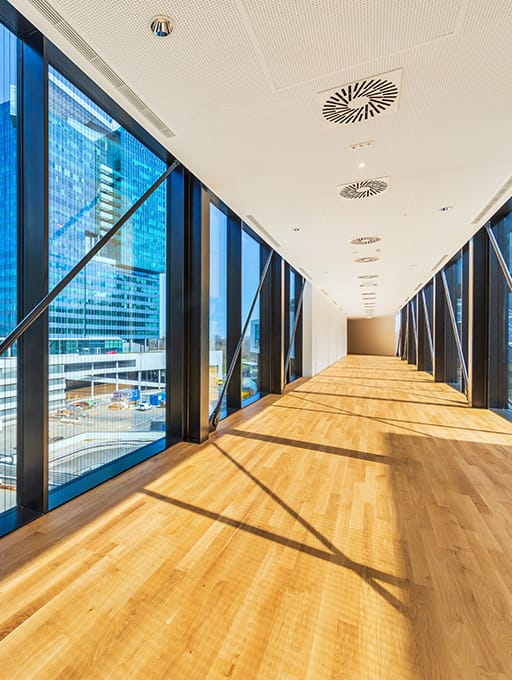 Langer Gang mit großen Fenstern, Holzboden und Deckenlüftungen, Tageslicht.