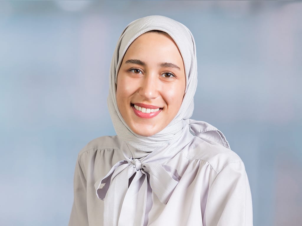 Person wearing a light-colored blouse with a bow and a matching headscarf against a plain background