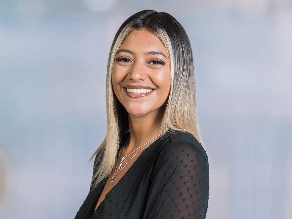 Person with long straight blonde hair wearing a black textured top and silver necklaces against a plain background