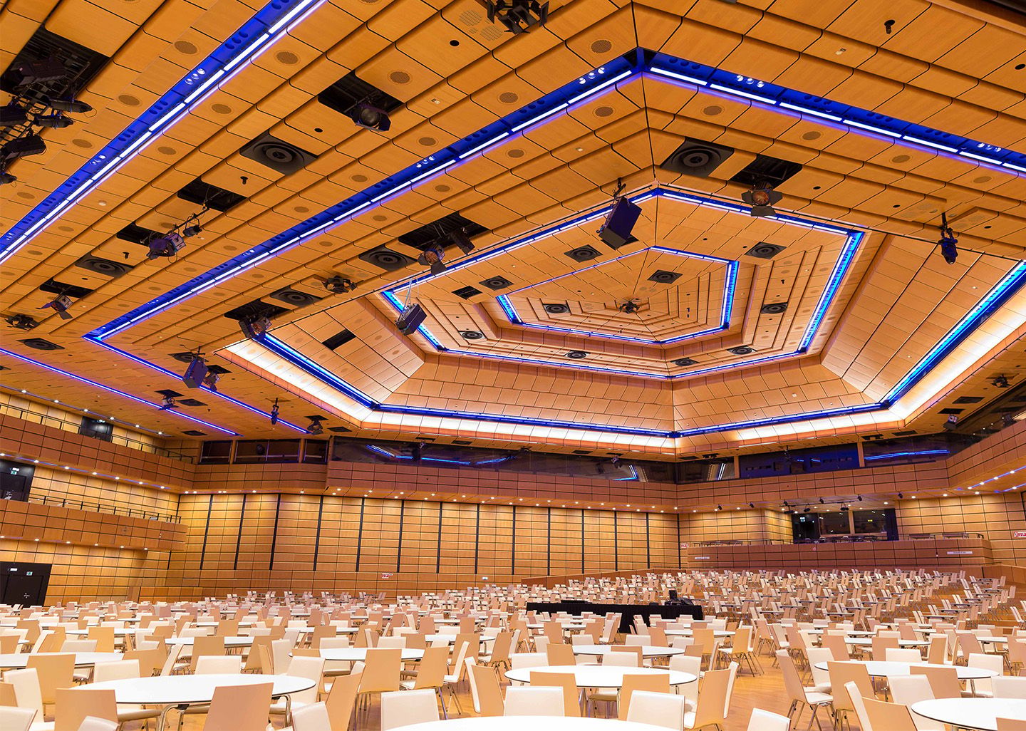 Large conference hall with round tables and beige chairs under a geometric ceiling with blue lights