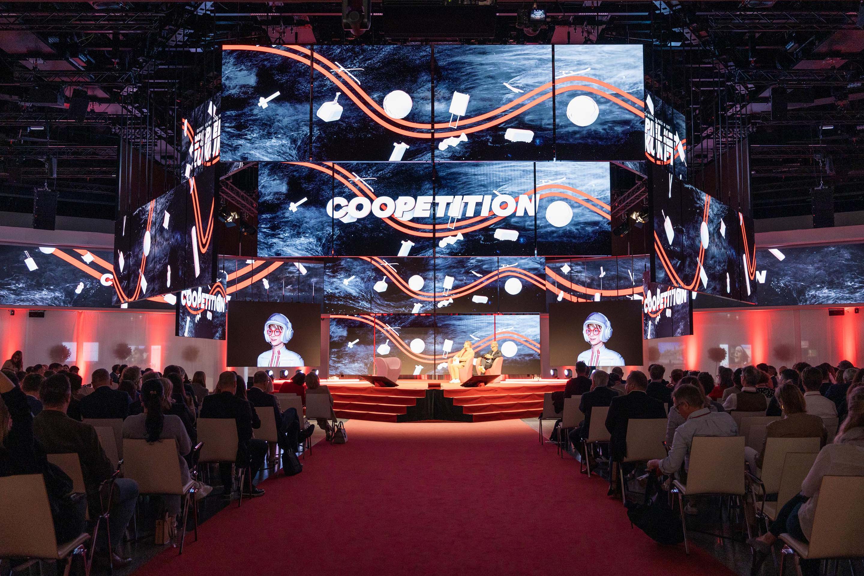 Konferenzsaal mit Publikum, Bühne und großen Bildschirmen mit dem Wort „COOPETITION“