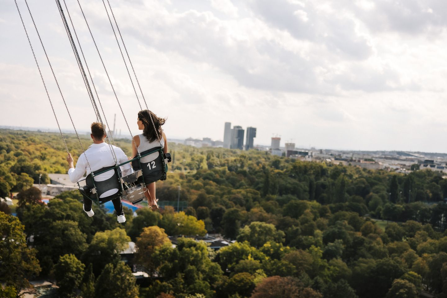 Two people sitting on swing ride above green trees with city skyline in distance