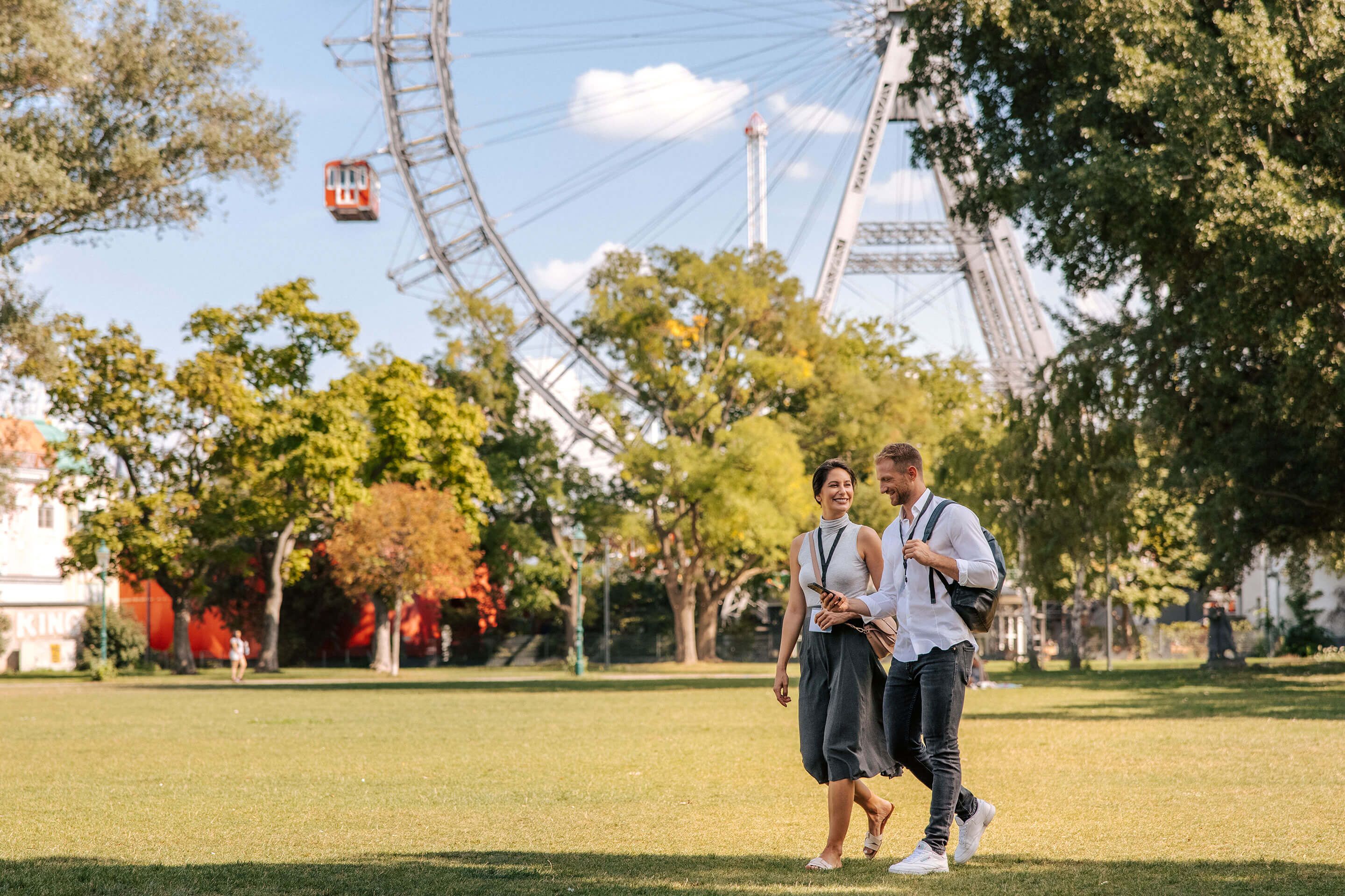Two people walking on grass near a large Ferris wheel and trees on a sunny day