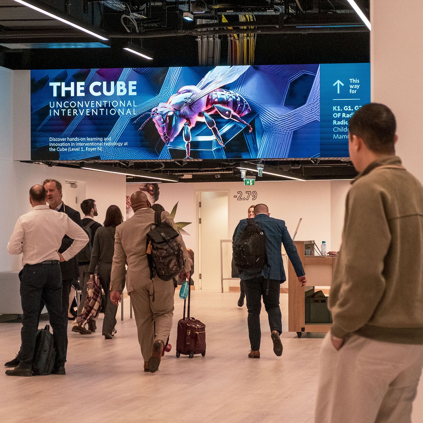 Menschen gehen in einem Flur unter einem Schild mit der Aufschrift „THE CUBE UNCONVENTIONAL INTERVENTIONAL“ und einem Bild einer Biene.