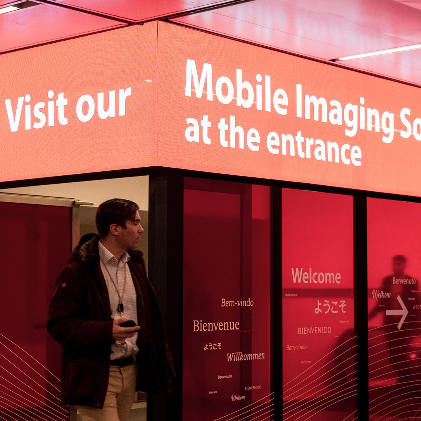 Person steht vor rotem Eingangsbereich mit mehrsprachigen Willkommensgrüßen und weißer Schrift "Mobile Imaging Social at the entrance"