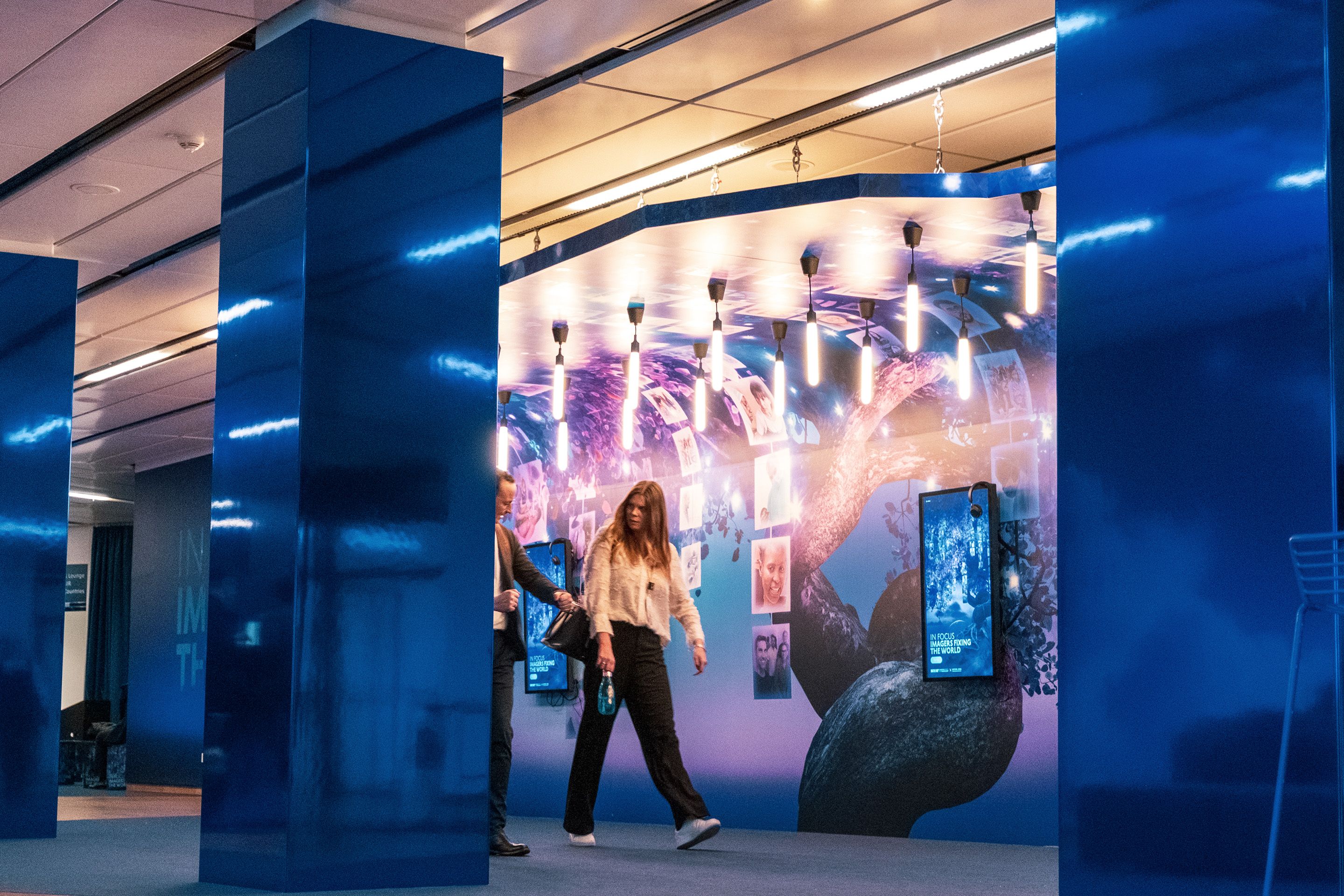 Two people walking past a blue-lit exhibition with hanging lights and digital displays.