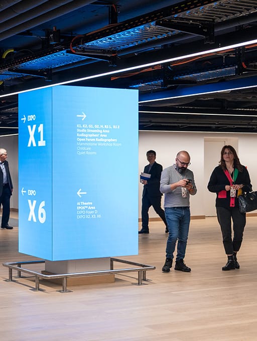 Leuchtender Wegweiser mit Hinweisen zu Expo X1 und X6 in einem Innenraum mit Personen.