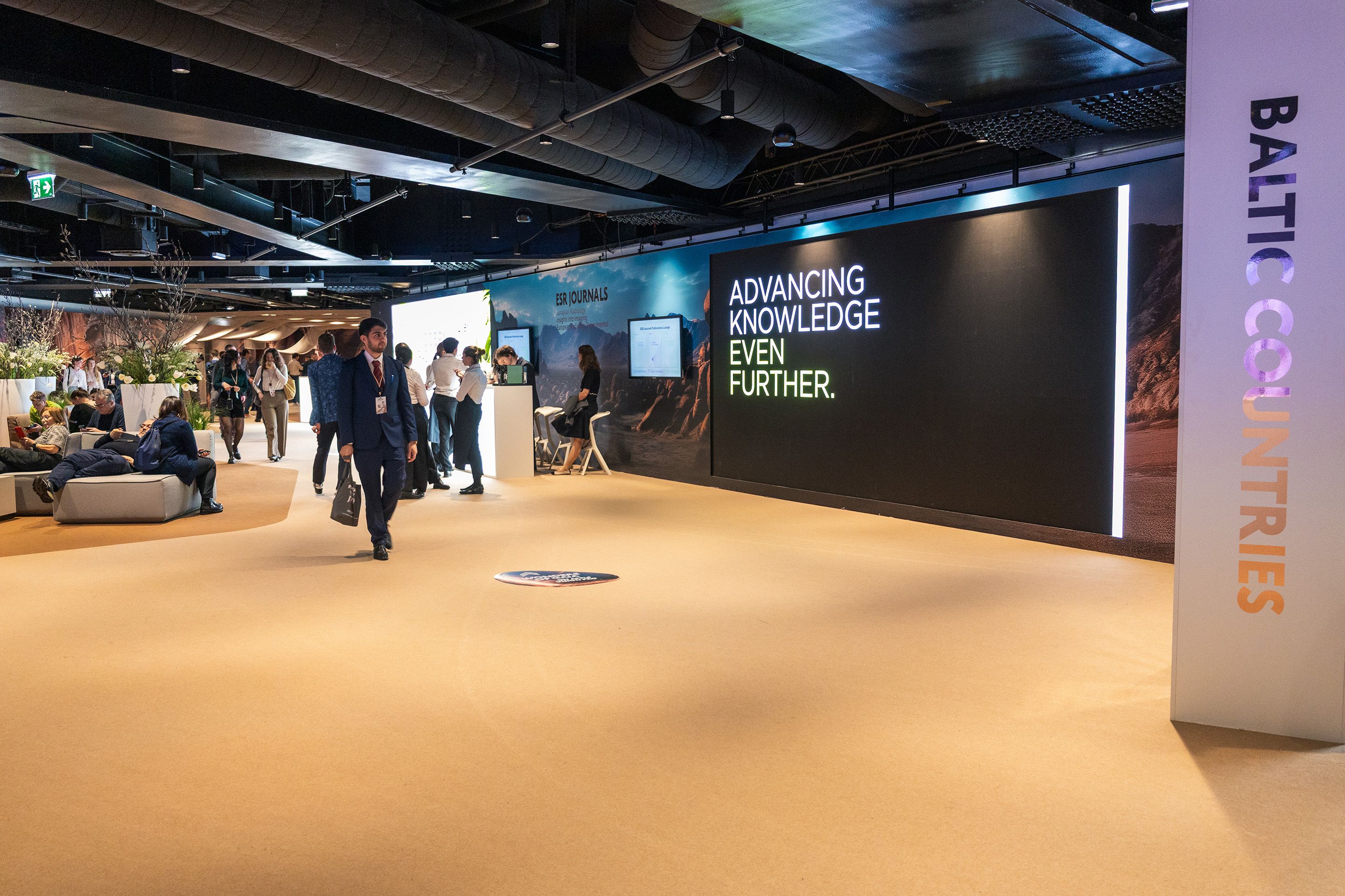 Messehalle mit Menschen, Sitzgelegenheiten und Leuchtschrift „ADVANCING KNOWLEDGE EVEN FURTHER.“