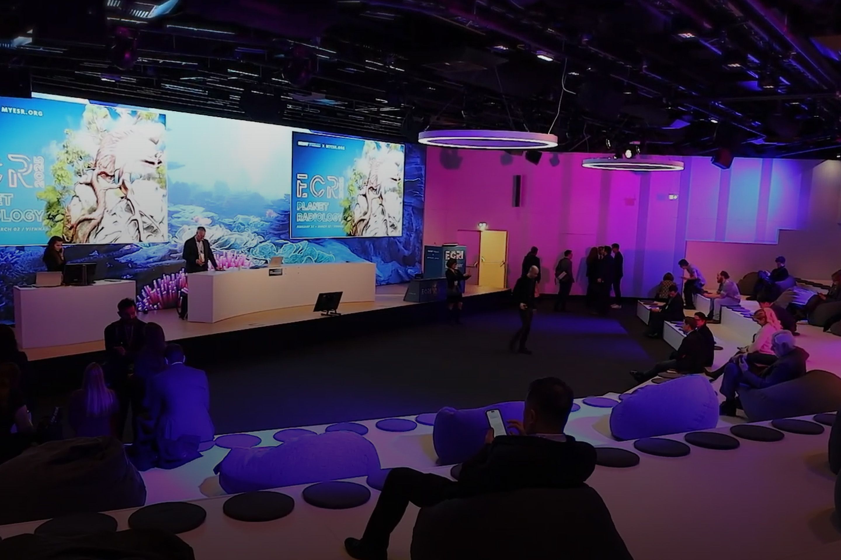 Conference room with stage, large screens displaying "ECR Planet Radiology," and seated audience.