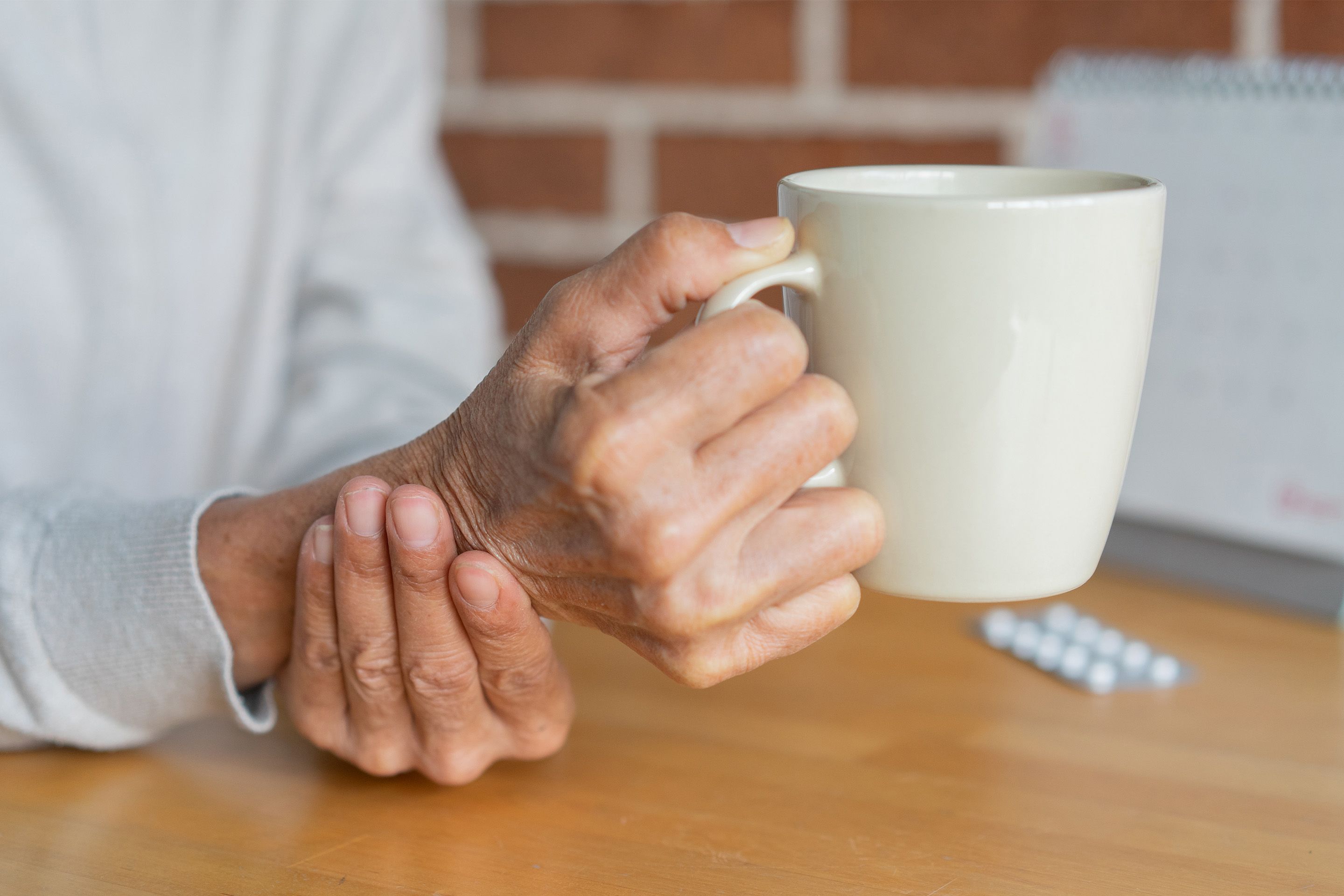 Ältere Hand hält Handgelenk, andere Hand hält weiße Tasse, Tablettenpackung auf Tisch