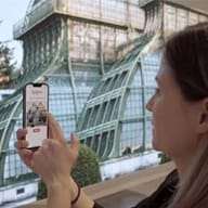 Person holding smartphone displaying a building with glass roof and metal framework in the background