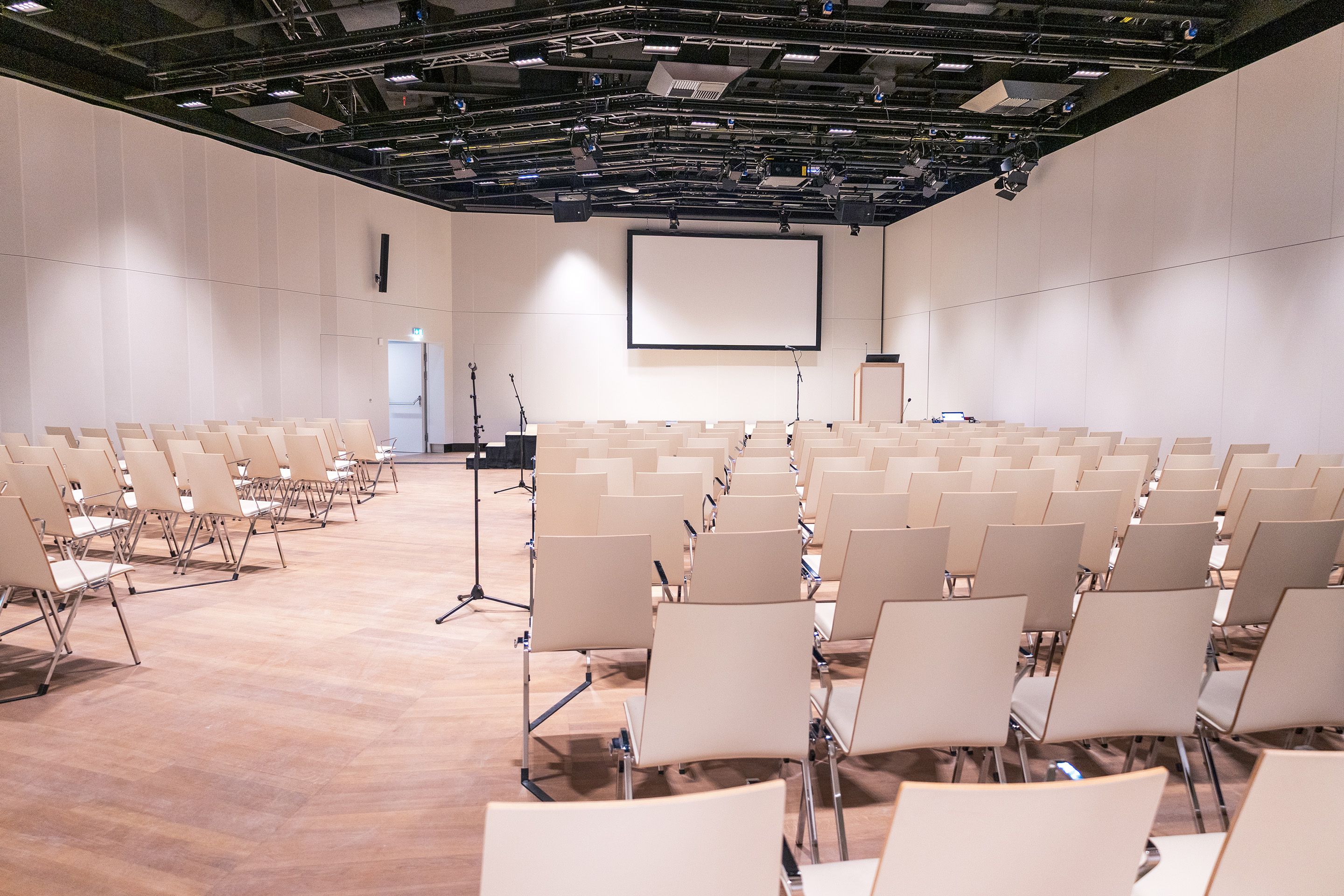 Leerer Konferenzraum mit Reihen von Stühlen, Mikrofonständern und Leinwand vorne.