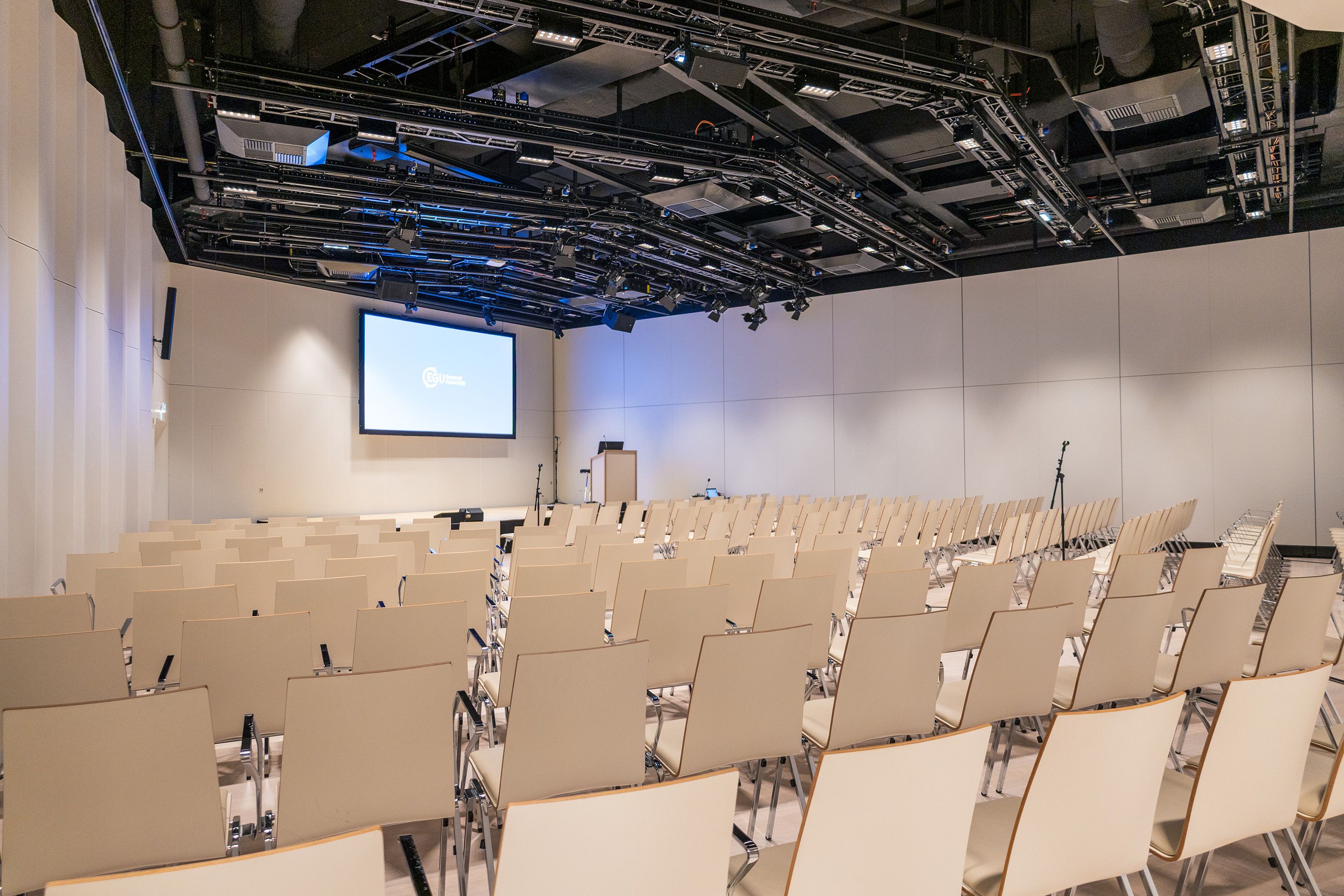 Leerer Konferenzraum mit Reihen von Stühlen, Leinwand und Mikrofonen auf der Bühne.