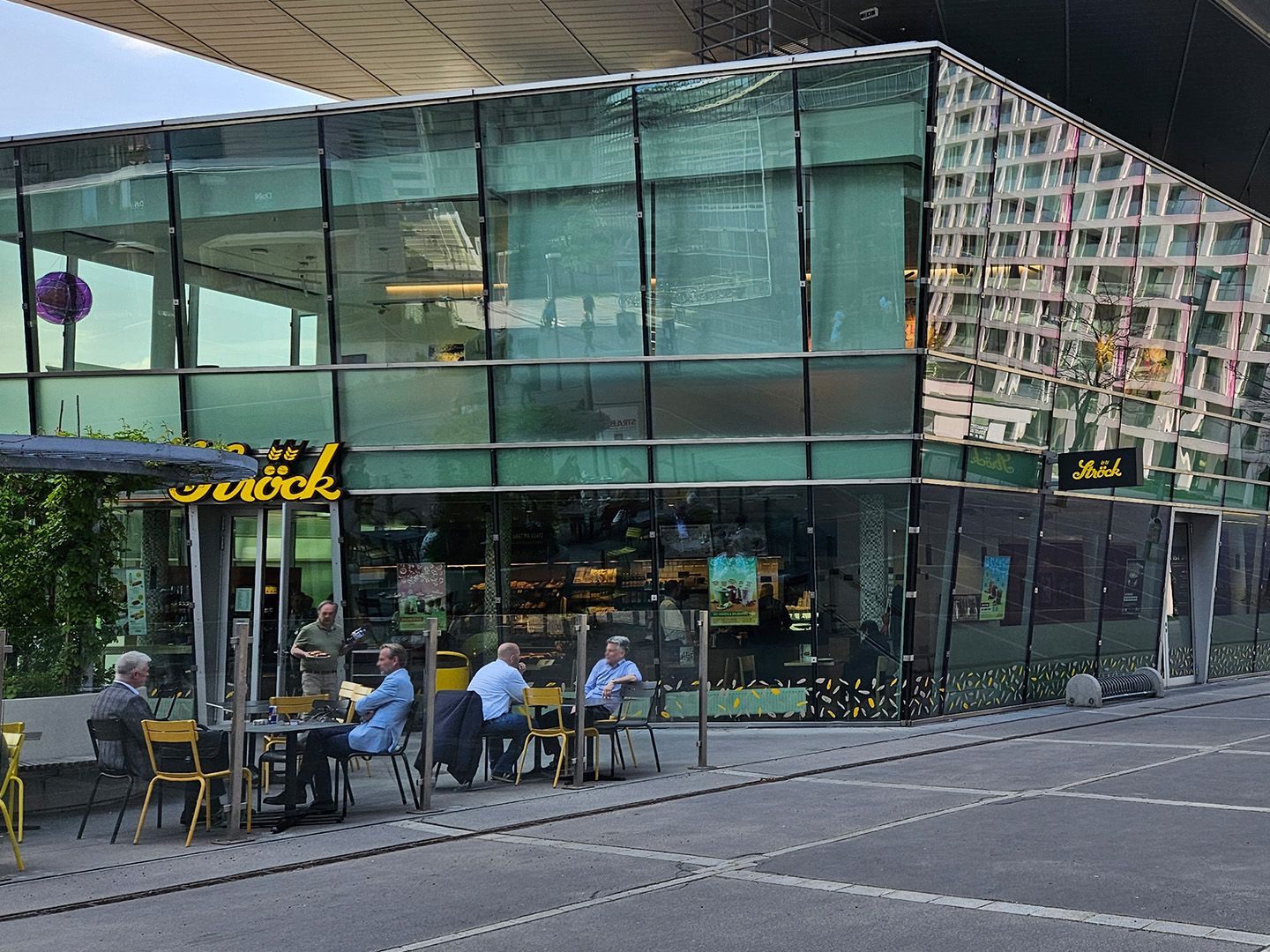 Außenbereich eines Cafés mit mehreren Personen an Tischen vor einem modernen Glasgebäude.