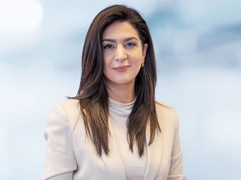 woman with long dark hair wearing a light beige blazer and white top against a blurred background