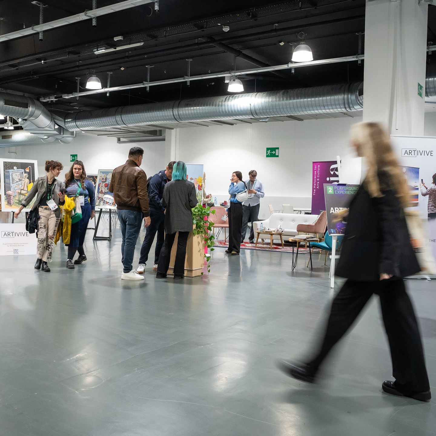 People walking and standing in a modern indoor exhibition space with art displays and seating areas.