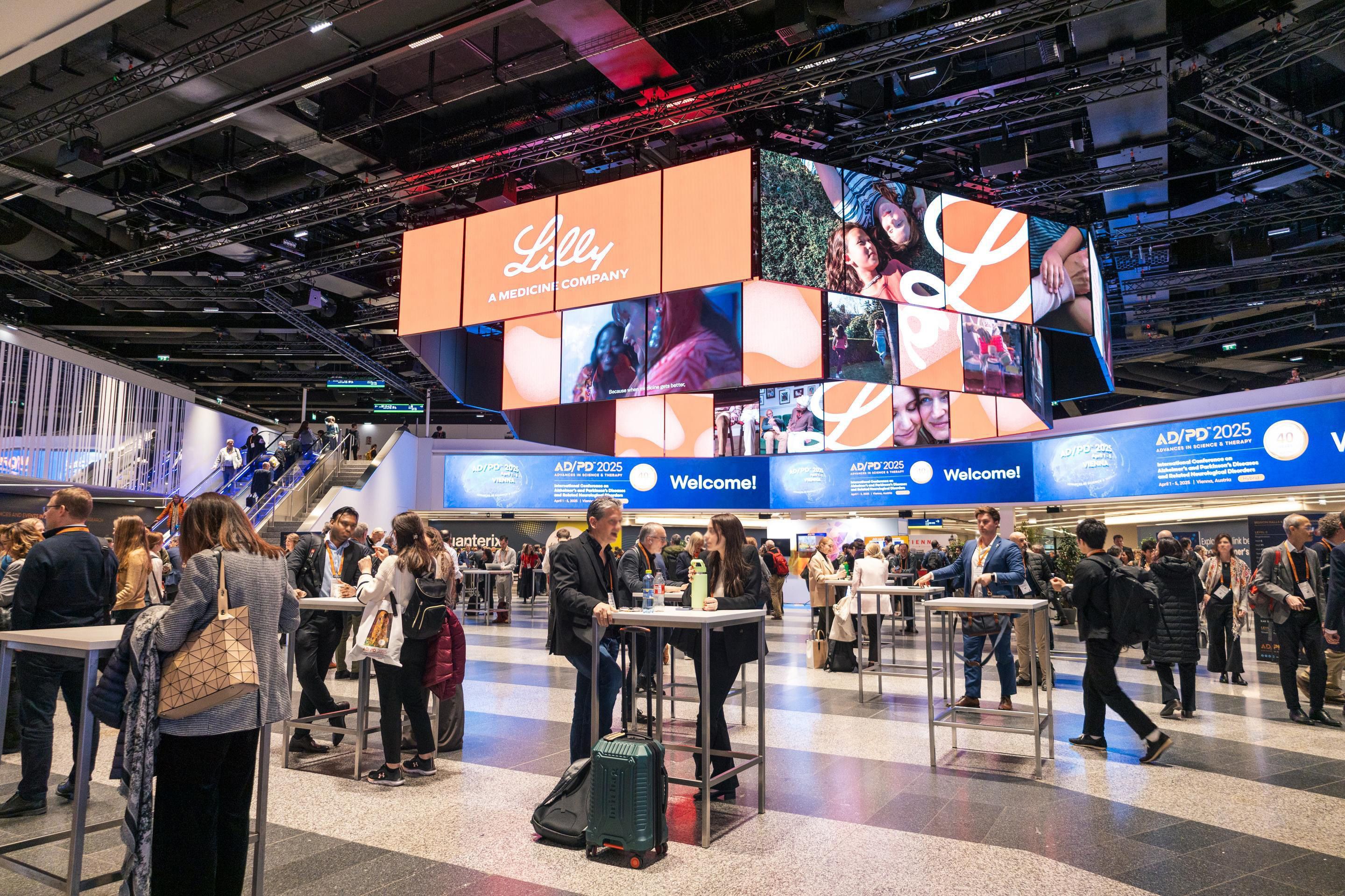 Menschen stehen an Stehtischen in einer großen Halle mit digitaler Werbewand und Rolltreppe.