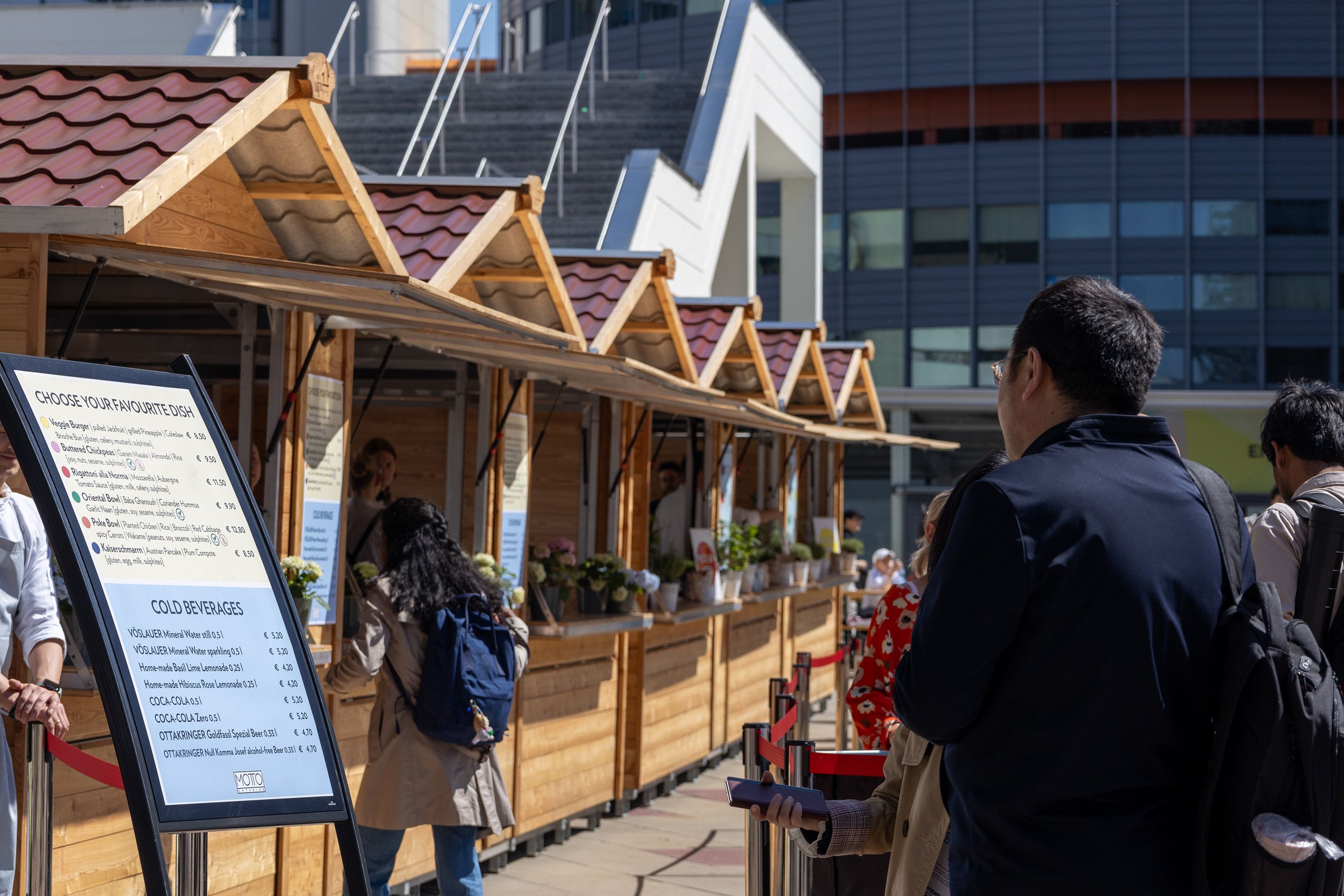 Outdoor food stalls with wooden roofs and a menu board listing dishes and cold beverages