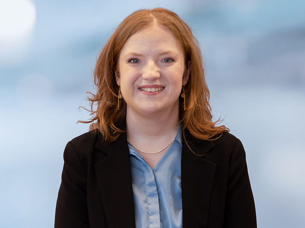 Person with shoulder-length red hair wearing a black blazer and light blue shirt against a blurred background