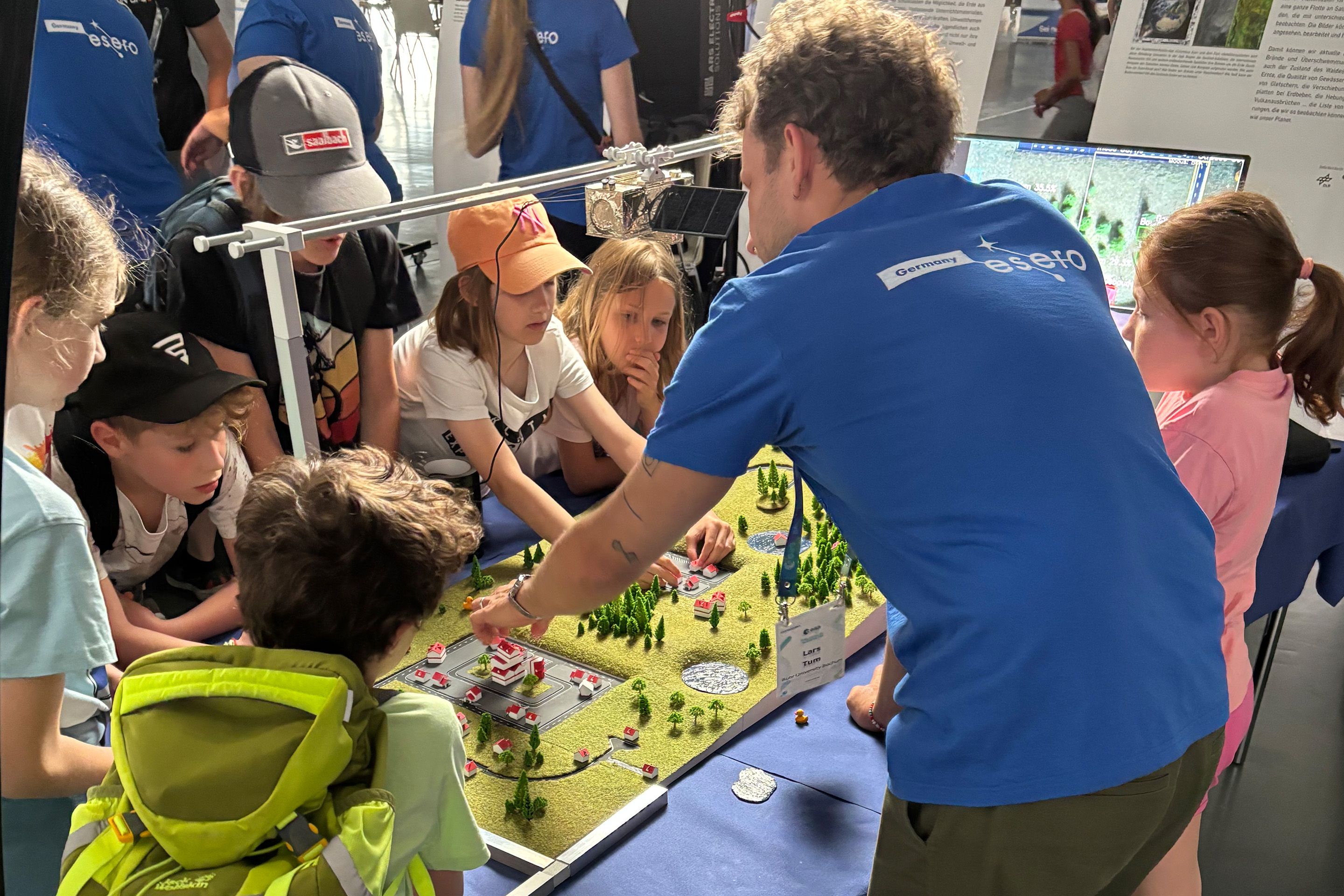 Person erklärt Kindern ein Modell mit Gebäuden, Bäumen und Straßen auf einem Tisch.