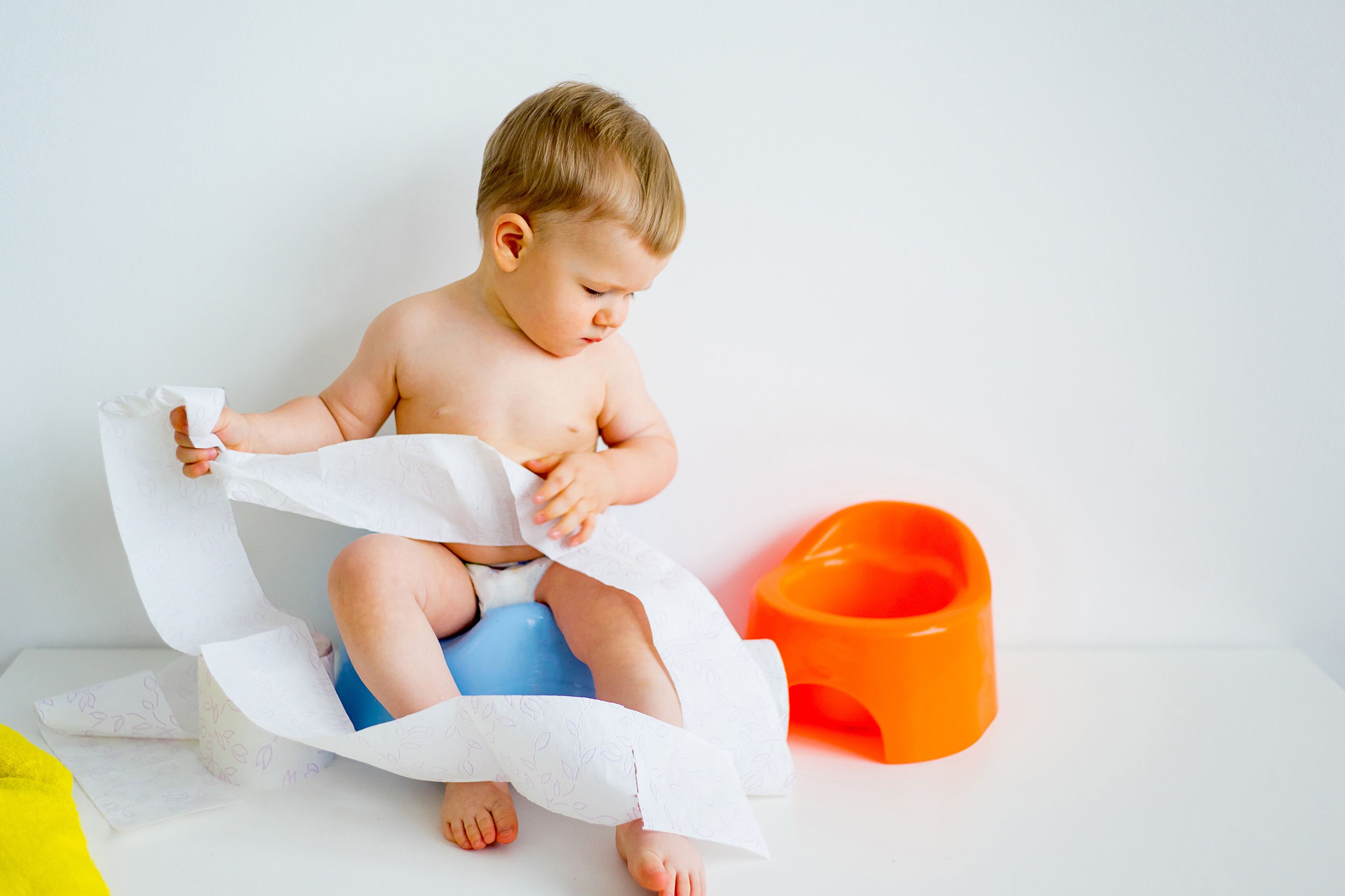 Kleinkind sitzt auf blauem Töpfchen und hält eine lange Papierrolle, daneben ein orangenes Töpfchen.