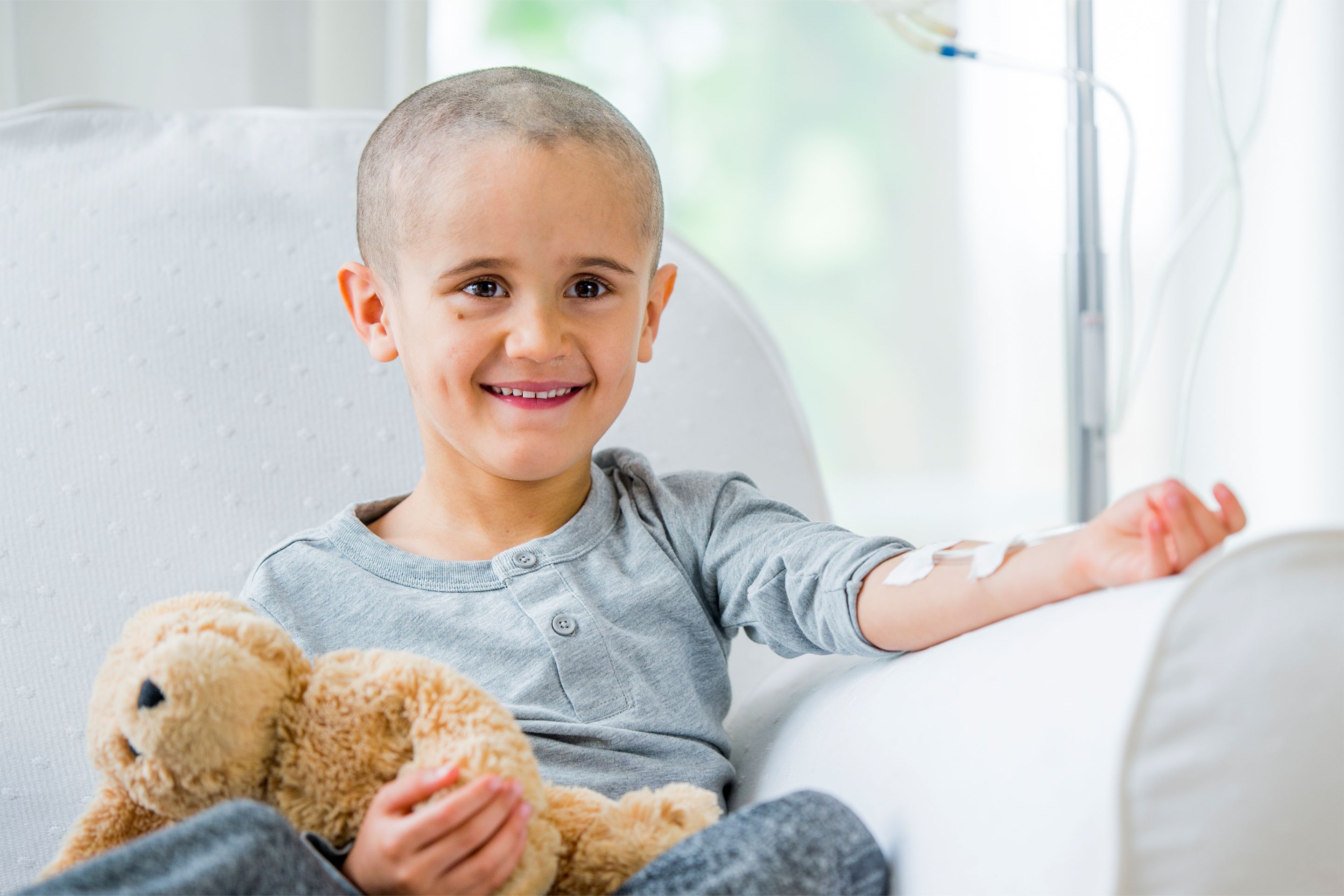 Kind mit Infusionskanüle am Arm sitzt auf Sofa und hält Teddybär.