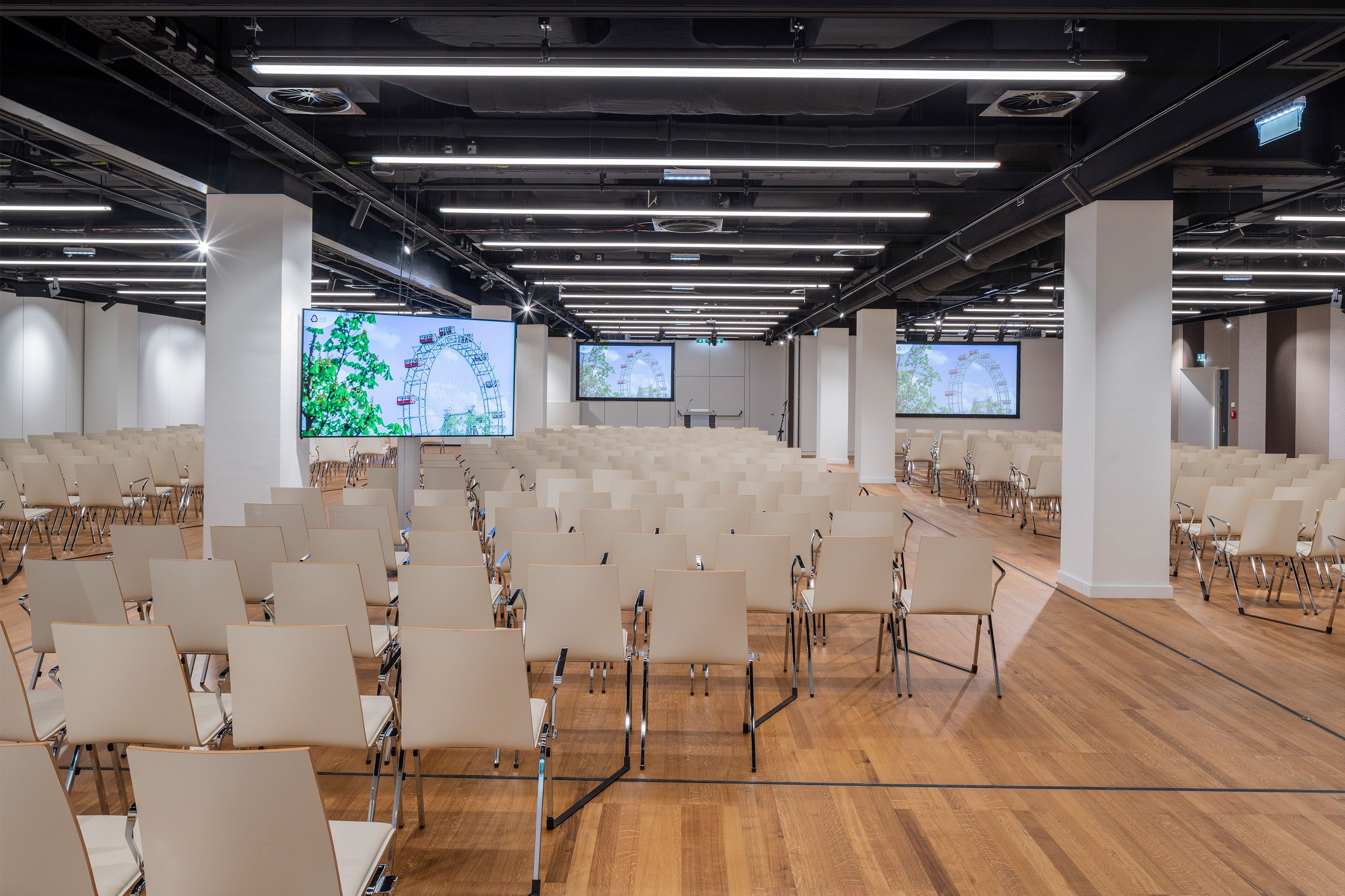 Foto: Sitzreihen in einem Saal mit Holzboden, Monitore, gestreifte Tapete