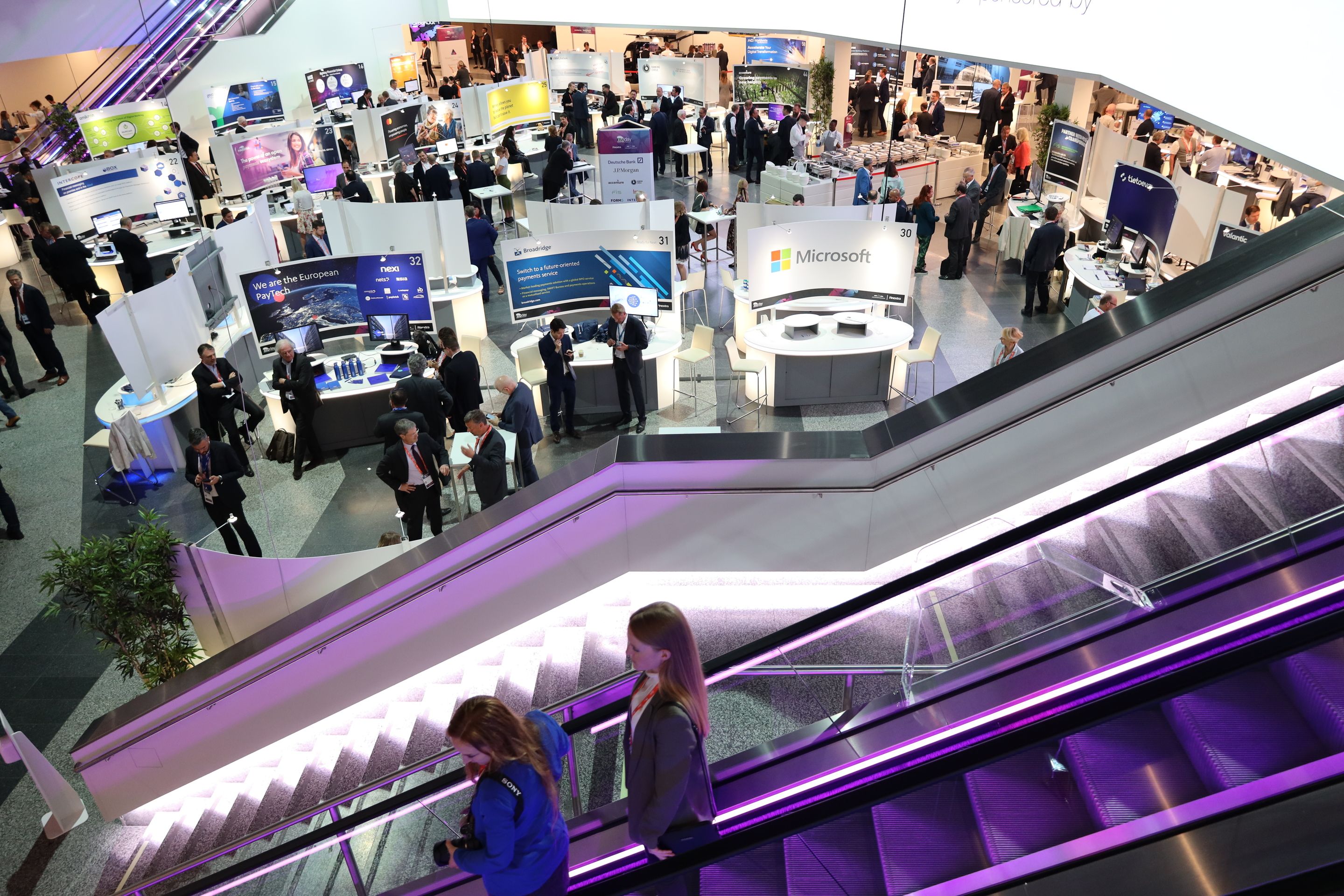Foto: lila beleuchtete Rolltreppe in einer Halle mit Ausstellern