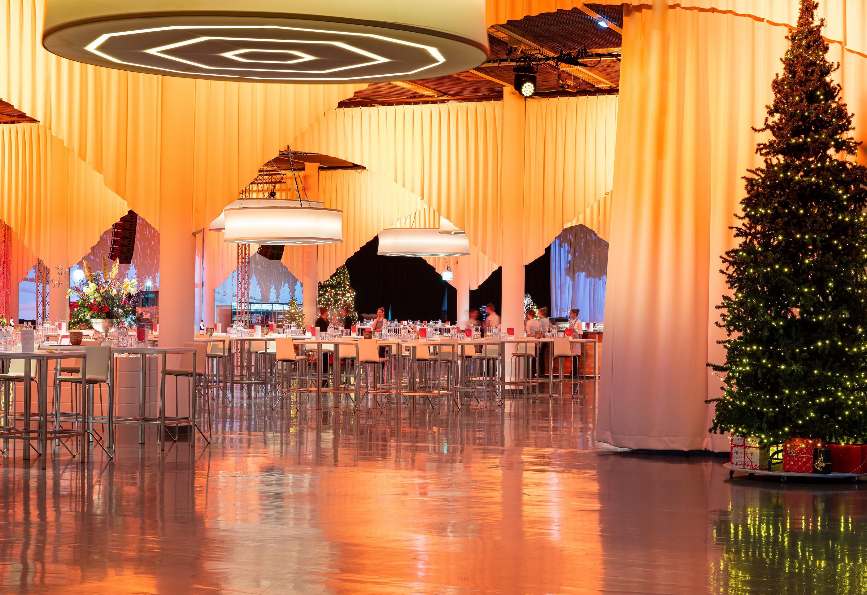 Foto: Eine Halle mit Christbaum, gedeckten Tischen, Lampeb und Vorhaengen