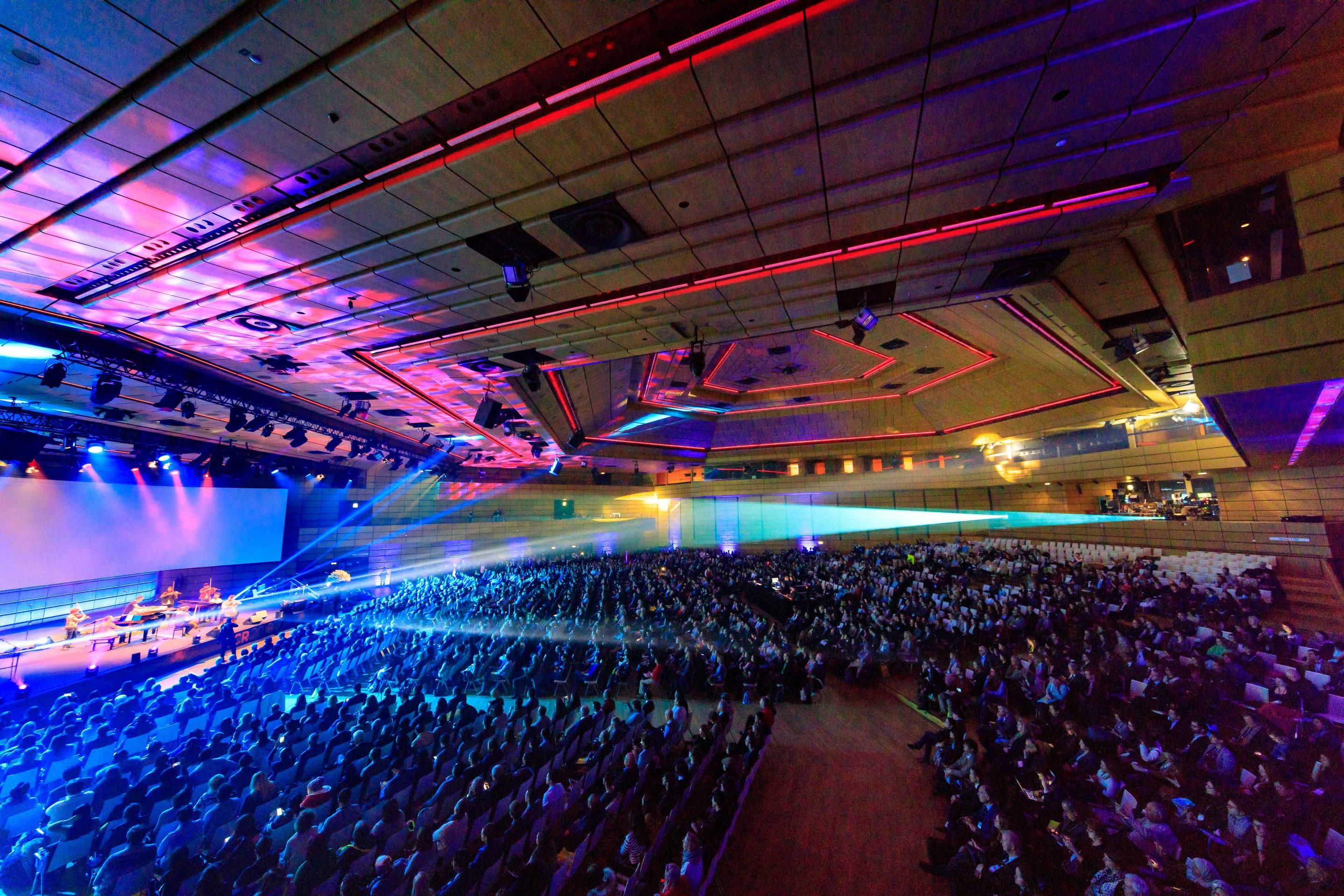 Foto: Halle voller Menschen sitzen vor einer Buehne, bunte Lichter
