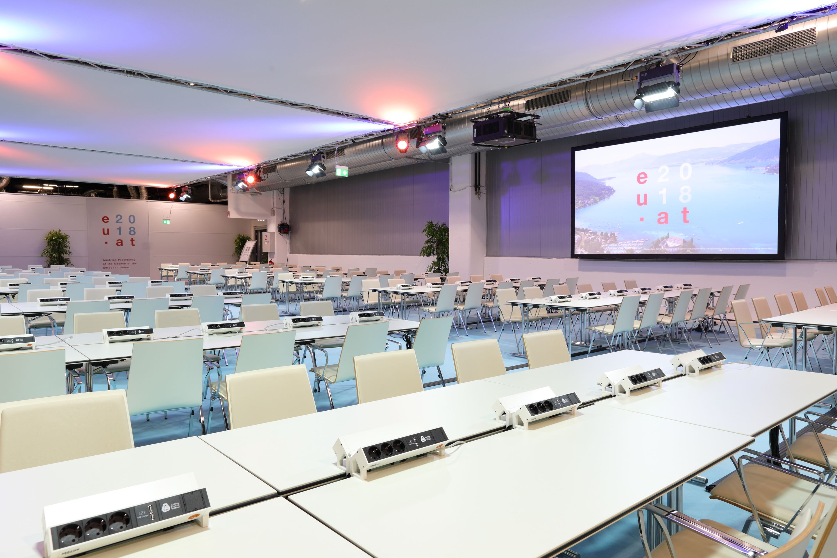 Foto: Weiße Tische und Sessel in einer Halle mit einer Leinwand