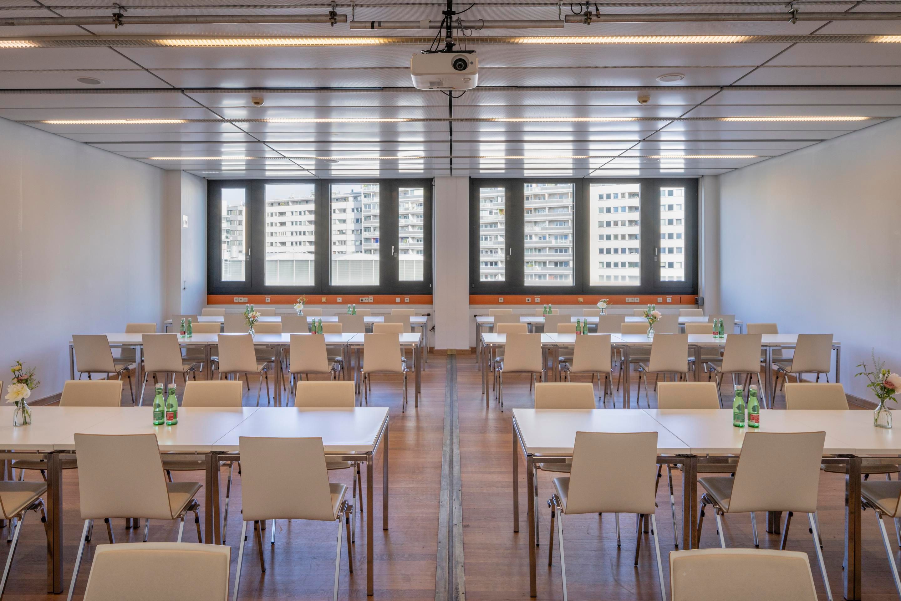 Foto: Raum mit Holzboden und Tischreihen mit Stuehlen. Durch das Fenster sieht man weisse Gebaeude.