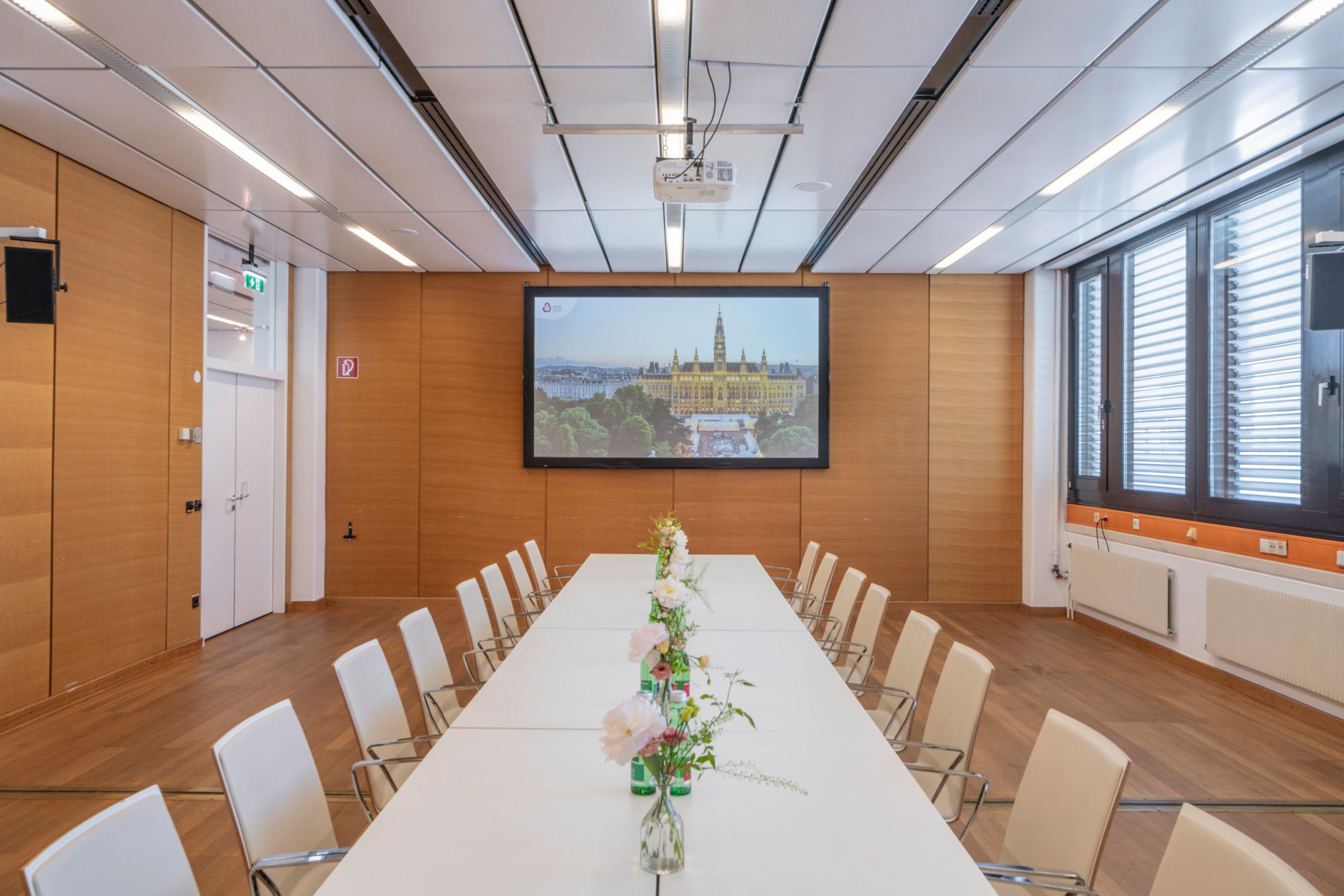 Foto: Raum mit Holzboden, Fenstern, weisse Tisch und Stuehlen. Eine Leinwand befindet sich an einer Holzwand.