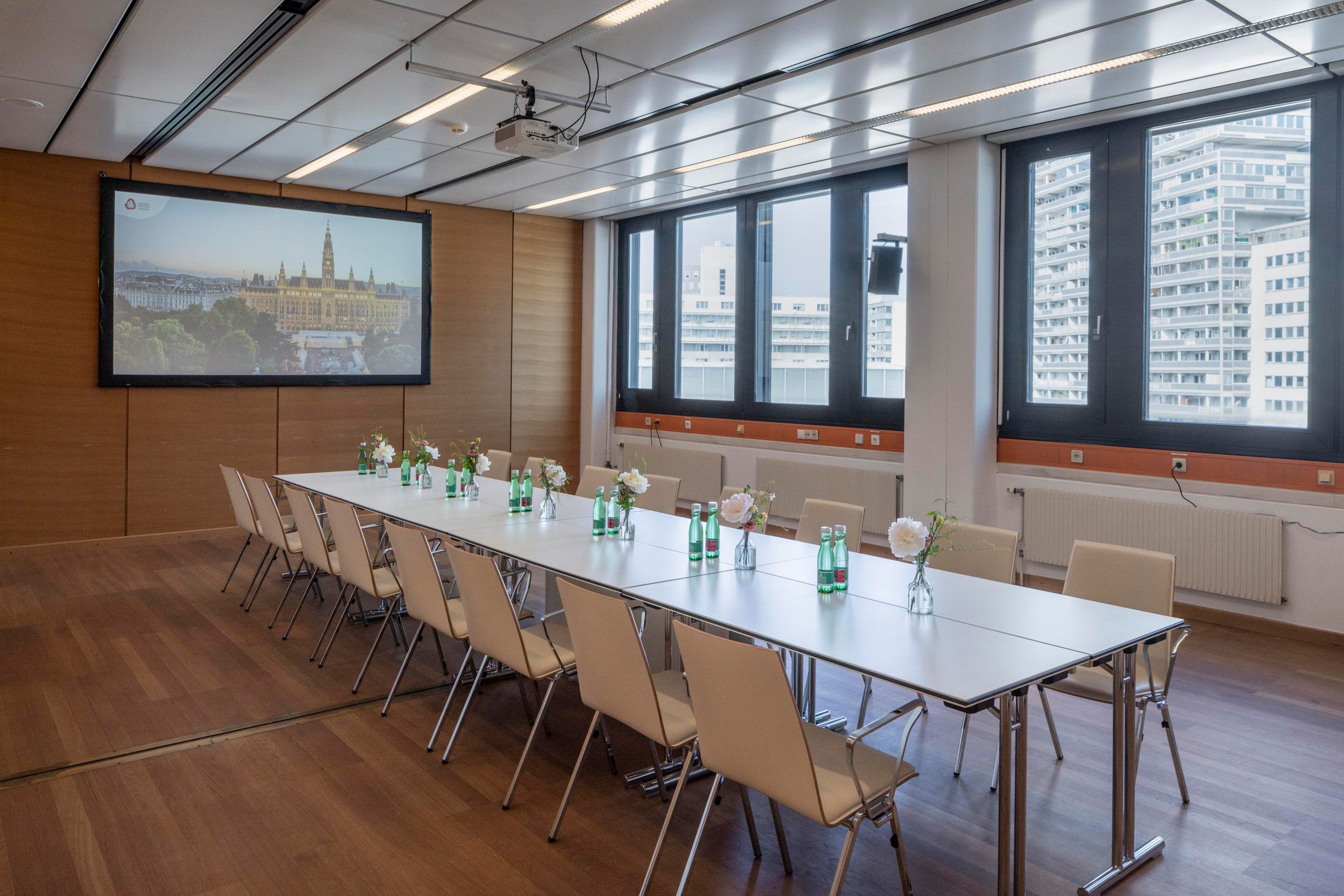 Foto: Raum mit Holzboden, Fenstern, weisse Tisch und Stuehlen. Eine Leinwand befindet sich an einer Holzwand.