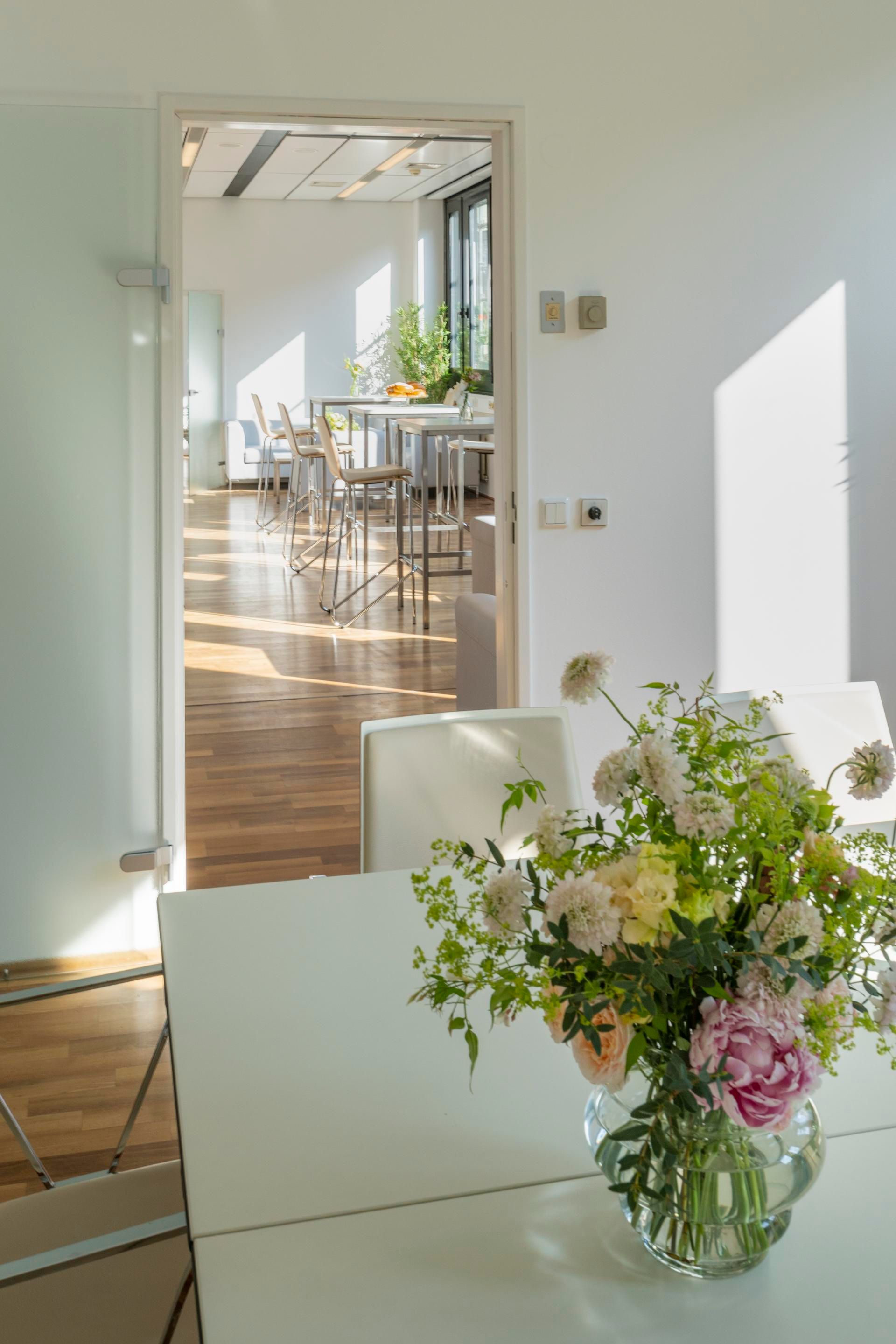 Foto: Raum mit Holzboden und einem hellen Tisch mit einer Blumenvase. Durch eine offene Tuer ist ein Nebenraum mit hohen Stuehlen und einem Tisch sichtbar.
