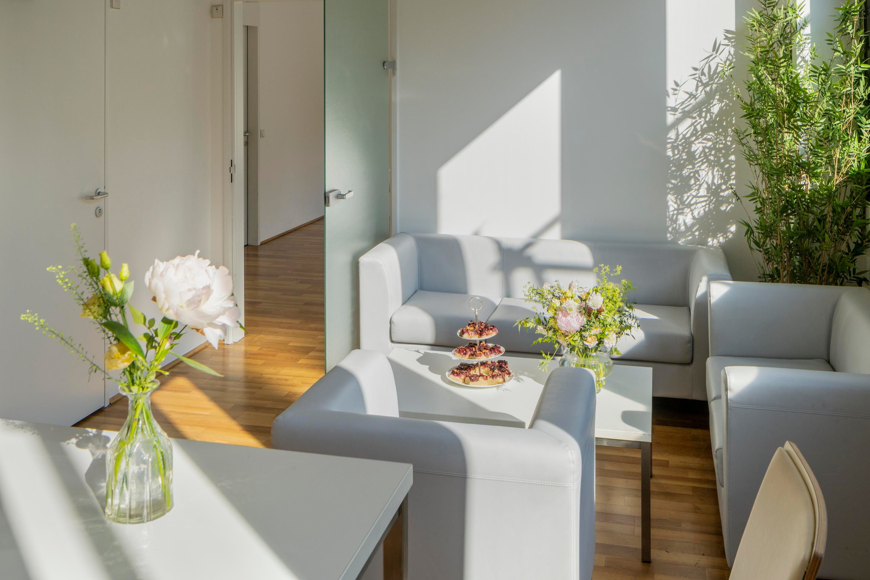 Foto: Raum mit Holzboden, hellen Sofas, Blumen und einer Etagere. Durch eine offene Tuer ist ein Nebenraum sichtbar.